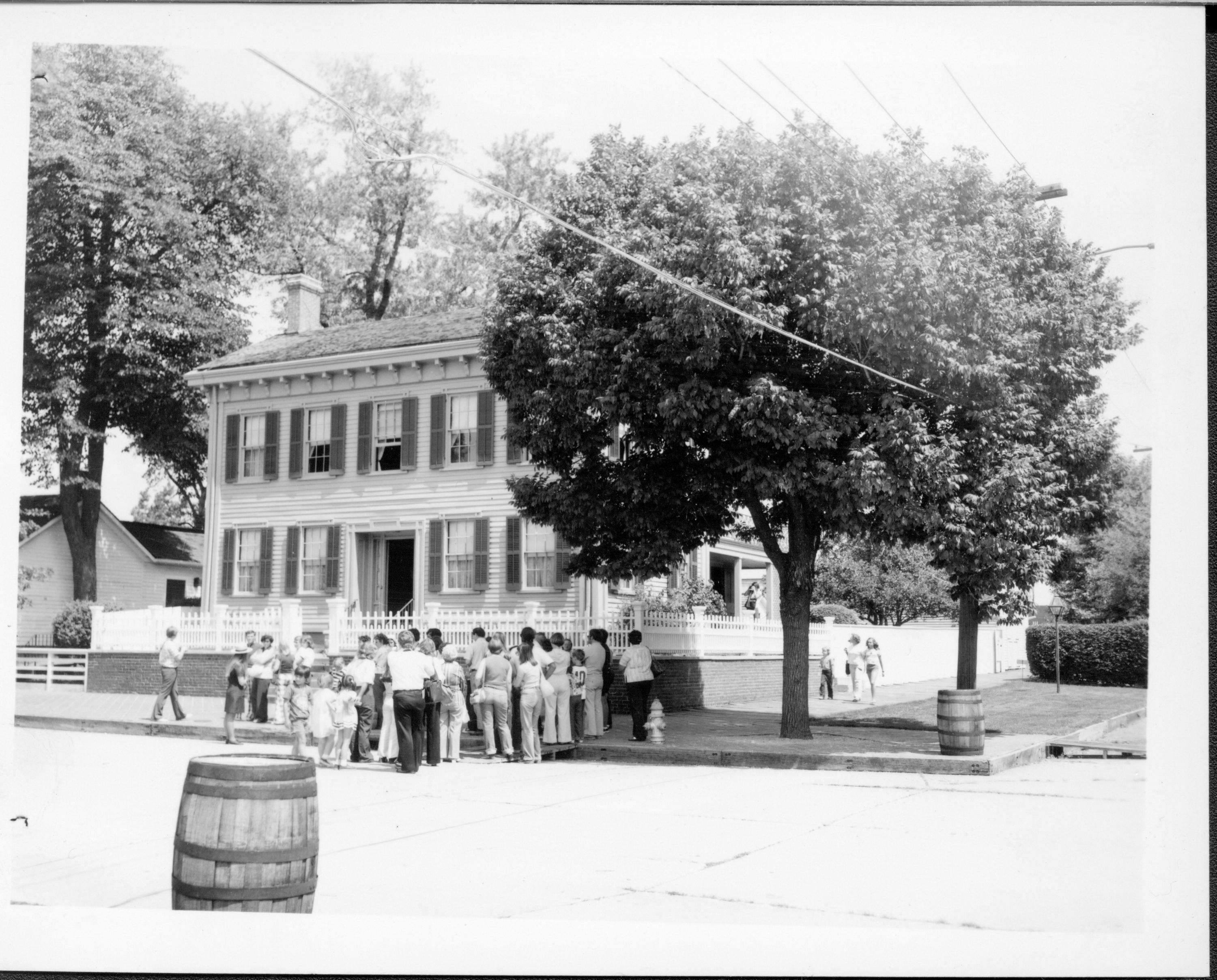 Lincoln Home front (West) elevation with visitors gathering in front of house listening to ranger in ranger hat on left standing in street. Other visitors visible on back porch and walking along boarwalk on south side of house. Trash barrels on corners near intersection. Fire hydrant on brick plaza near corner of house. Corneau House on far left. Electrical lines seen overhead.   Looking Northeast from 8th and Jackson Street intersection Lincoln Home, visitors, staff, Corneau, trash barrels, fire hydrant, 8th, Jackson