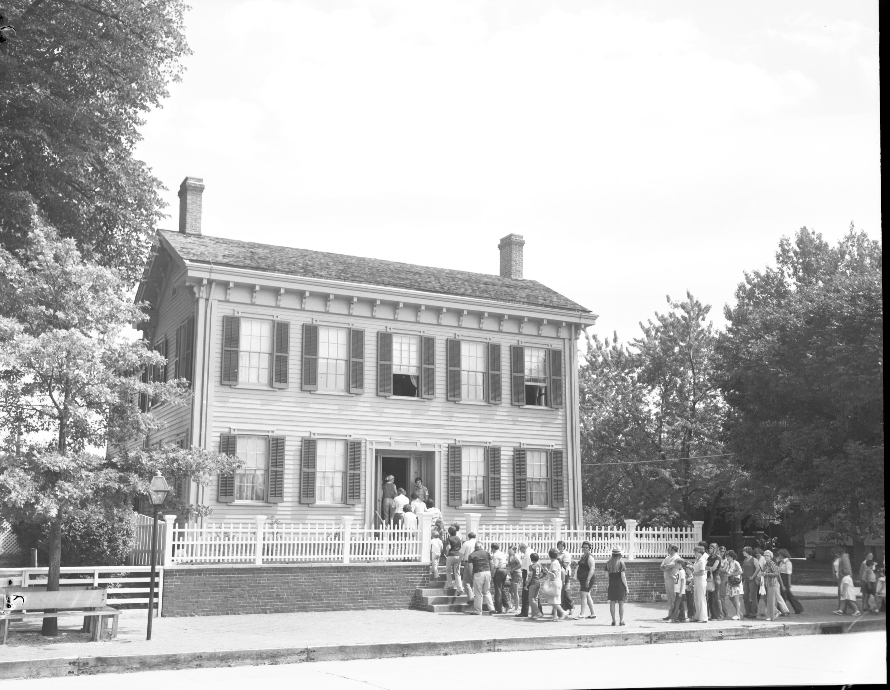 Lincoln Home front (West) elevation with visitors lining up to go inside.  A ranger in ranger hat is visible in line-break on brick plaza. Another ranger with no hat is standing to the right of the open front door. Window are open on the second floor in the center and far right. A bench circles the oak tree on far left. The Arnold House (dark) and Rebecca Cook houses are in far background right Looking Southeast from 8th Street Lincoln Home, visitors, staff, Arnold, Rebecca Cook, 8th Street