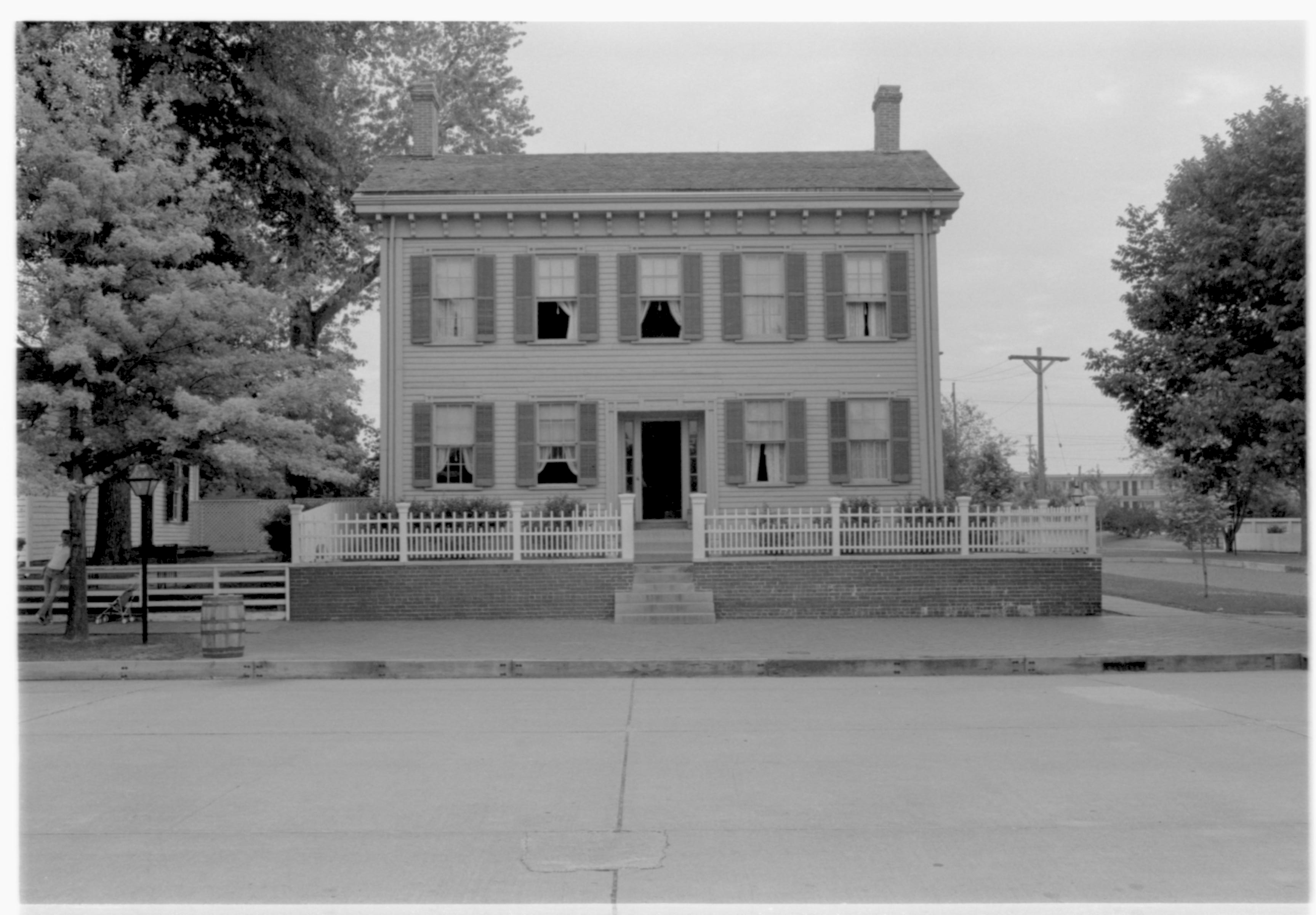 Lincoln Home front (West) elevation. Windows open on both floors. Visitors and a baby stroller visible on far left in front of Corneau House.  Electrical poles in background right. Travel Lodge Motel visible in far background right. Trash barrel on edge of brick plaza on left.  Looking East from West of 8th Street. Lincoln Home, 8th, Corneau, visitors, Travel Lodge Motel, electric poles