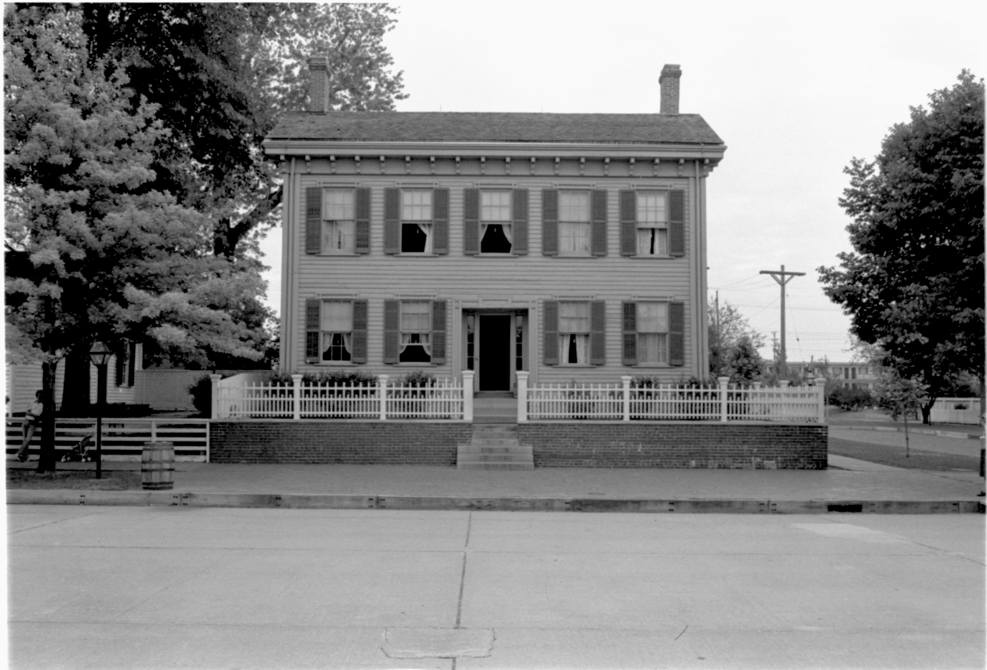 Lincoln Home front (West) elelvation. Visitors and a baby stroller visible on far left in front of Corneau House. Electrical pole in background right, Travel Lodge Motel in far background right. Ranger Ruth Ketchum just visible on far right. Trash barrel on edge of brick plaza on left. Looking East from West side of 8th Street Lincoln Home, visitors, staff, Corneau, Travel Lodge Motel, 8th, trash barrel