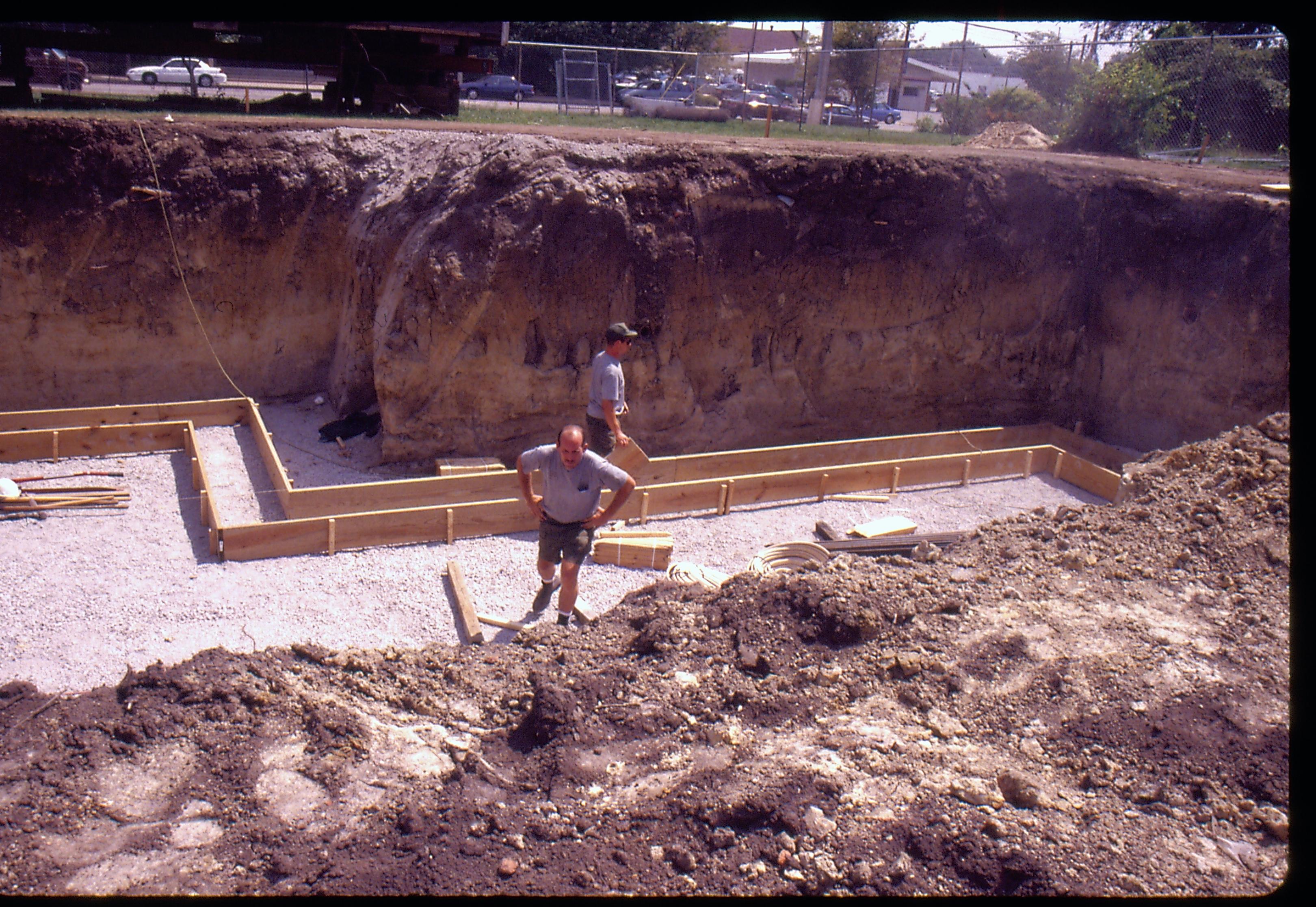 Morse - basement excavation, Setting wall footings. Maintenance workers Al McHenry and Jeff Daly? check footings before the concrete pour. 9th Street traffic visible in background. Looking East/Southeast from Morse Lot (Block 10, Lot 15-16) Morse, foundation, footings, staff, 9th Street