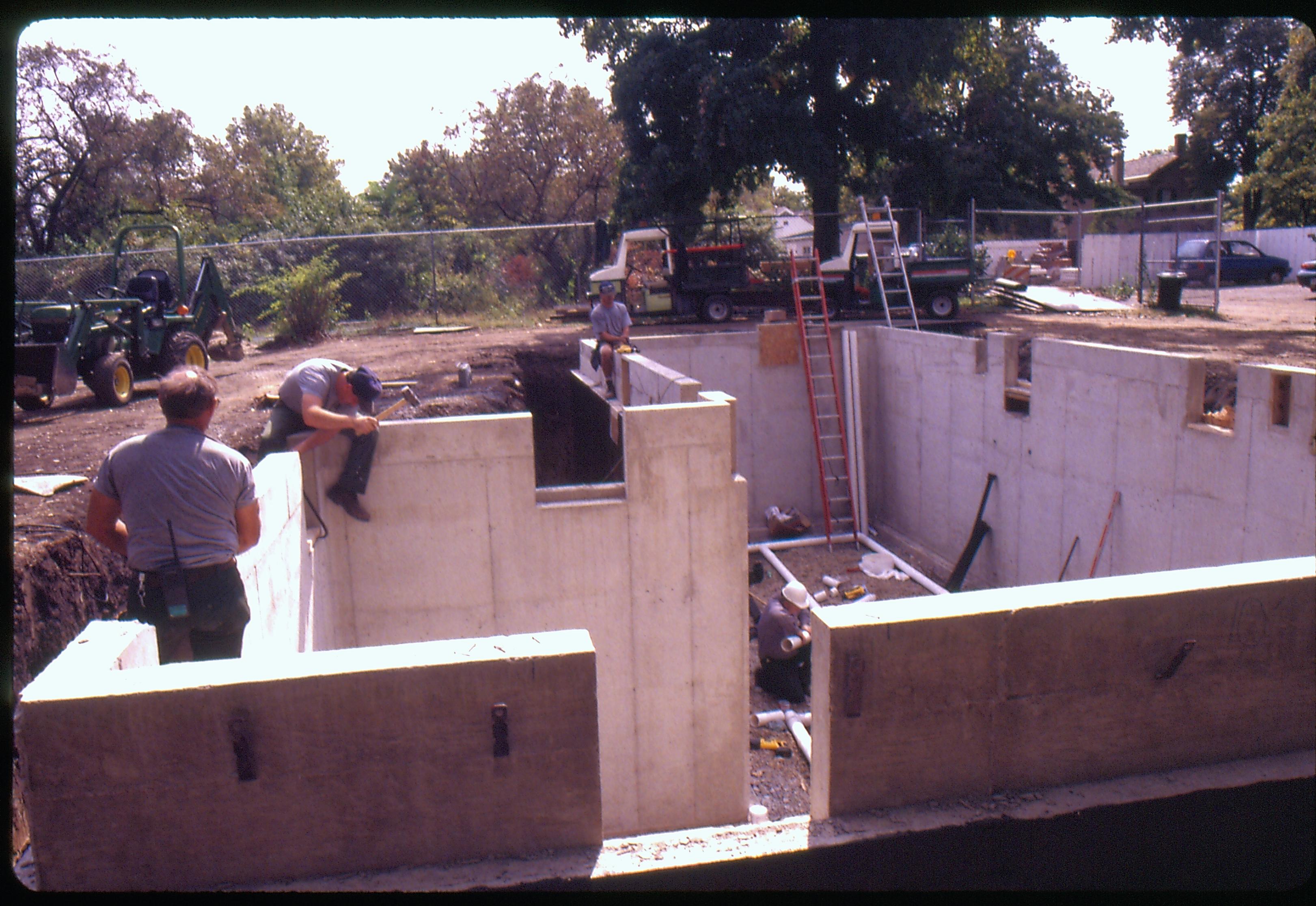 Morse - basement excavation, Walls poured. Maintenance Workers Sam Blasingame and ? and ? break out forms. Maintenance worker Tom Melton? lays pipe on floor.  Cushmans and backhoe in background. Looking South from Morse Lot (Block 10, Lots 15-16) Morse, foundation, walls, staff, cushman, backhoe