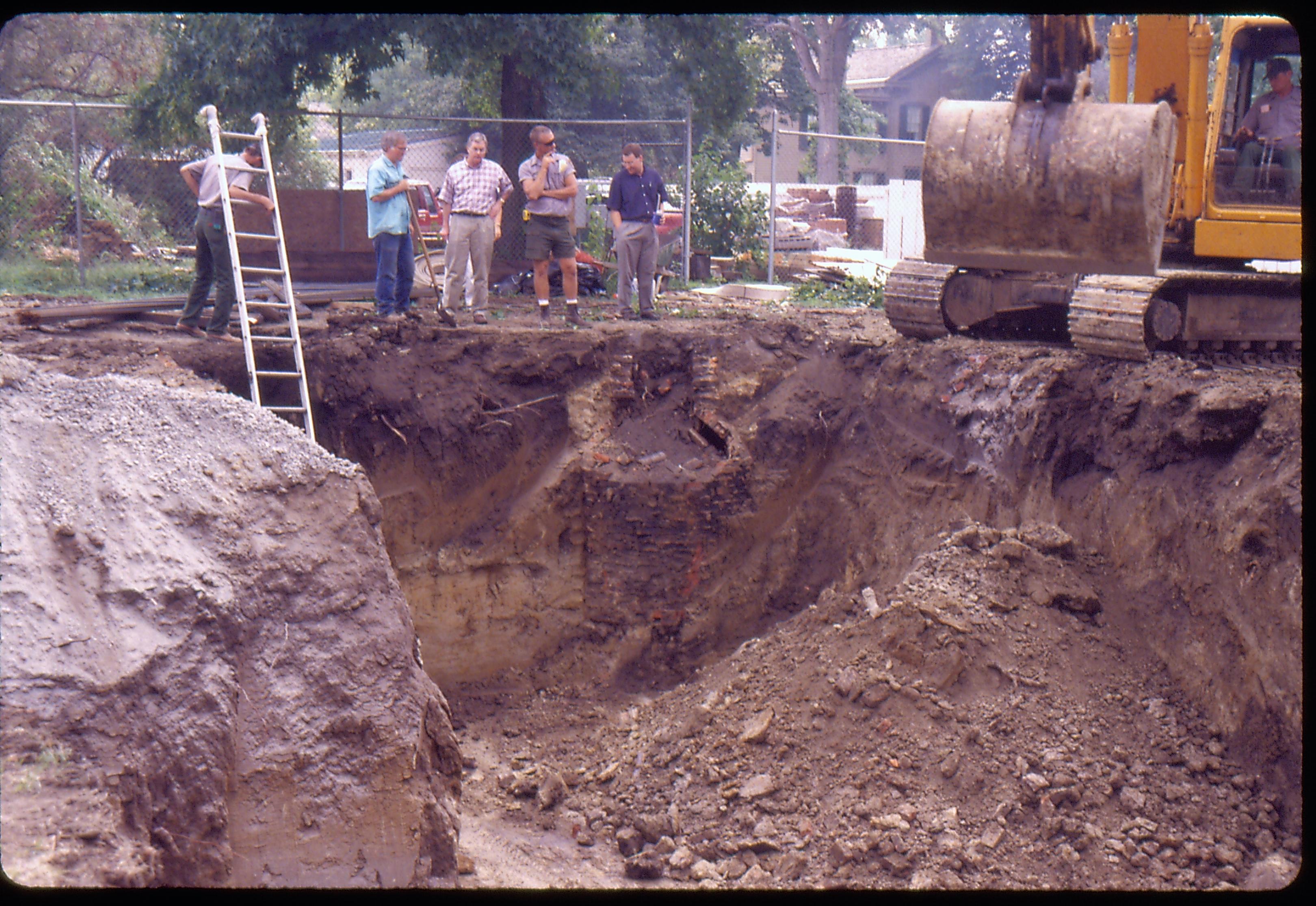 Morse - Basement excavation (cistern found). Maintenance worker Doug Sharp leans on ladder, Fever River Archeologist Floyd Mansberger (with shovel), Supt. Norman Hellmers, Maintenance Worker Ed Smith and Historian Tim Townsend watch as Maintenance Worker Vee Pollack works back hoe. Brick cistern is revealed in center.  Lincoln Home in background right. B-2 in background center left. Looking South/Southeast from Morse lot (Block 10, Lots 15-16) Morse, foundation, cistern, archeology, staff, Lincoln Home, B-2, backhoe, Fever River