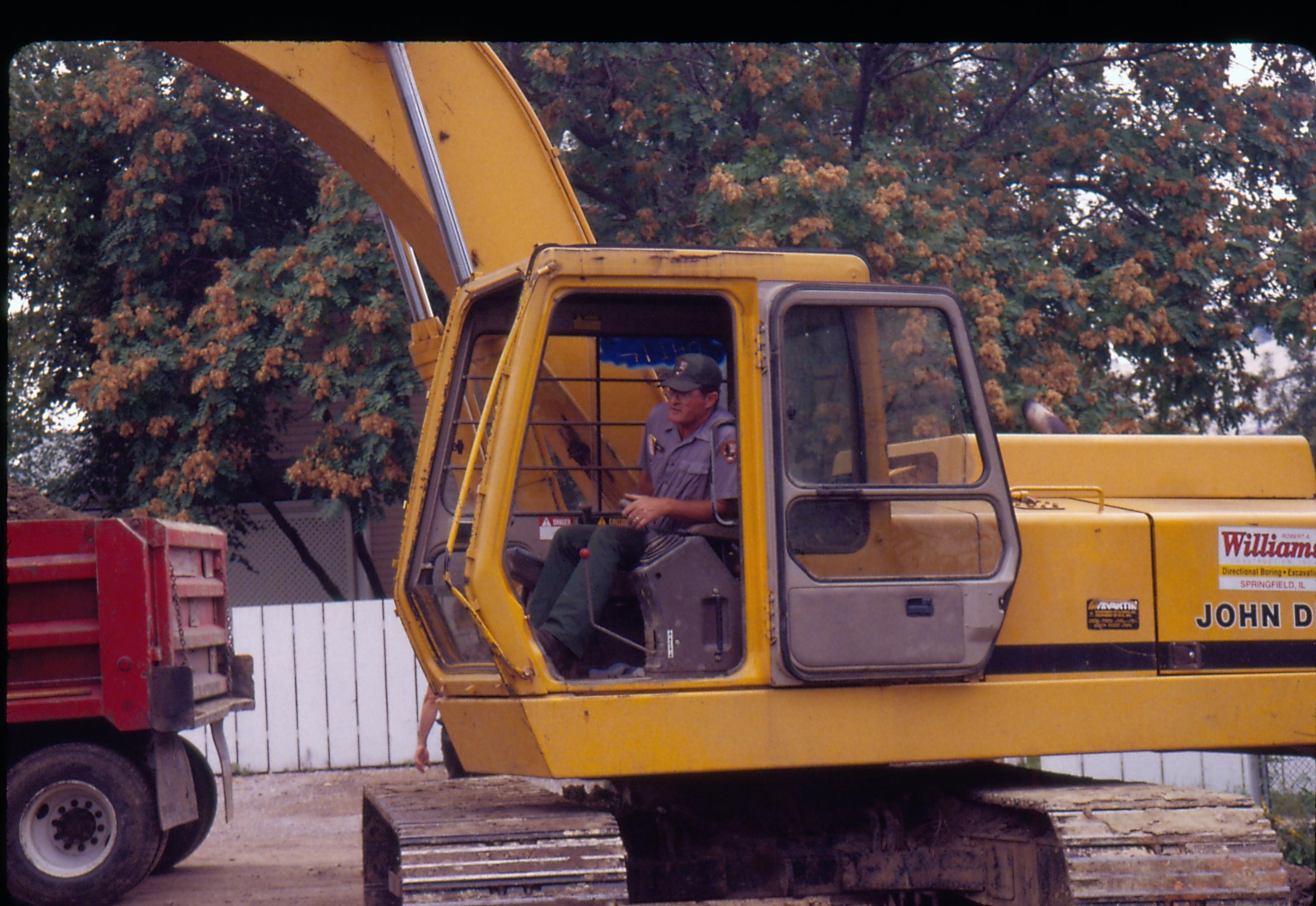 Morse - baseament excavation. Maintenance Worker Vee Pollack operating backhoe while waste hauler truck waits in alley. Latticework from Conference Center in background center. Looking West from Morse Lot (Block 10, Lots 15-16) Morse, foundation, Conference Center, backhoe, staff