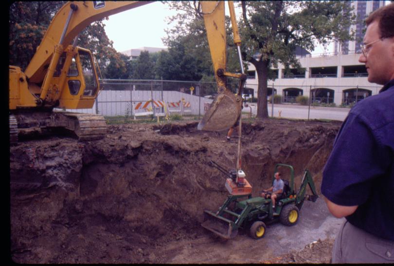 Morse - basement excavation with backhoe on edge of pit lowering equipment into pit.  Maintenance worker Al McHenry on smaller backhoe in pit. Maintenance Work Ed Smith's legs visible behind large backhoe bucket. Historian Tim Townsend in foreground Looking Northwest from Morse Lot (Block 10, Lots 15-16) Morse, foundation, excavation, backhoe, staff