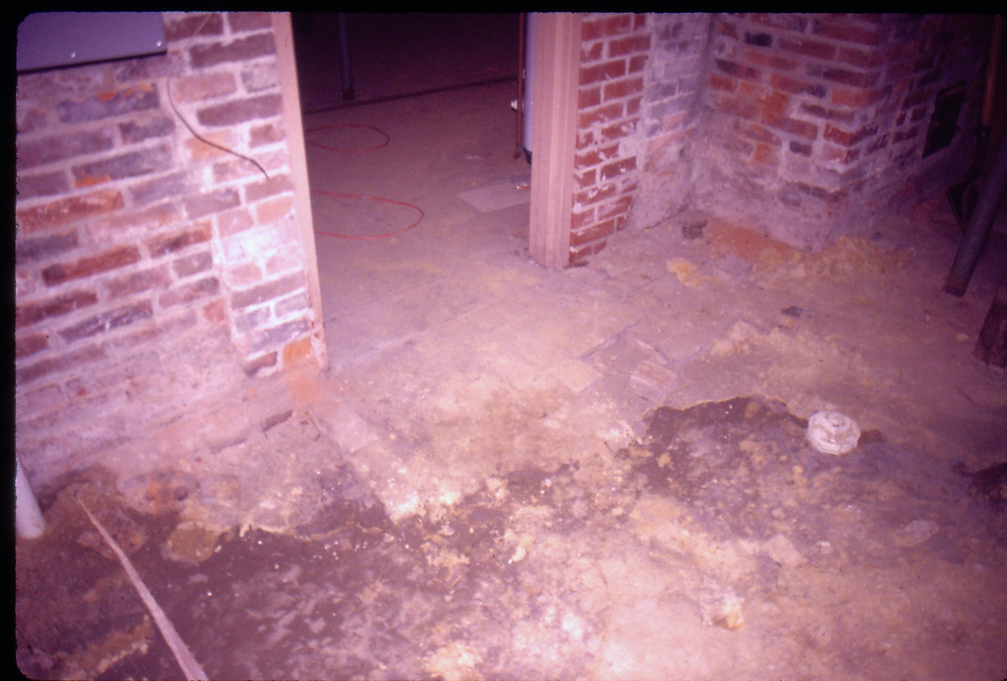 Lyon House - basement, floor and wall leading into furnace room in background. Floor is mix of dirt and old brick in a pattern, white capped drain on right. Brick walls visible with doorway leading to furnace room.  Furnace room appears to have concrete floor. Extension cord visible on floor in furnace room Looking Northwest from basement Lyon, Basement, brick floor, brick wall, furnace