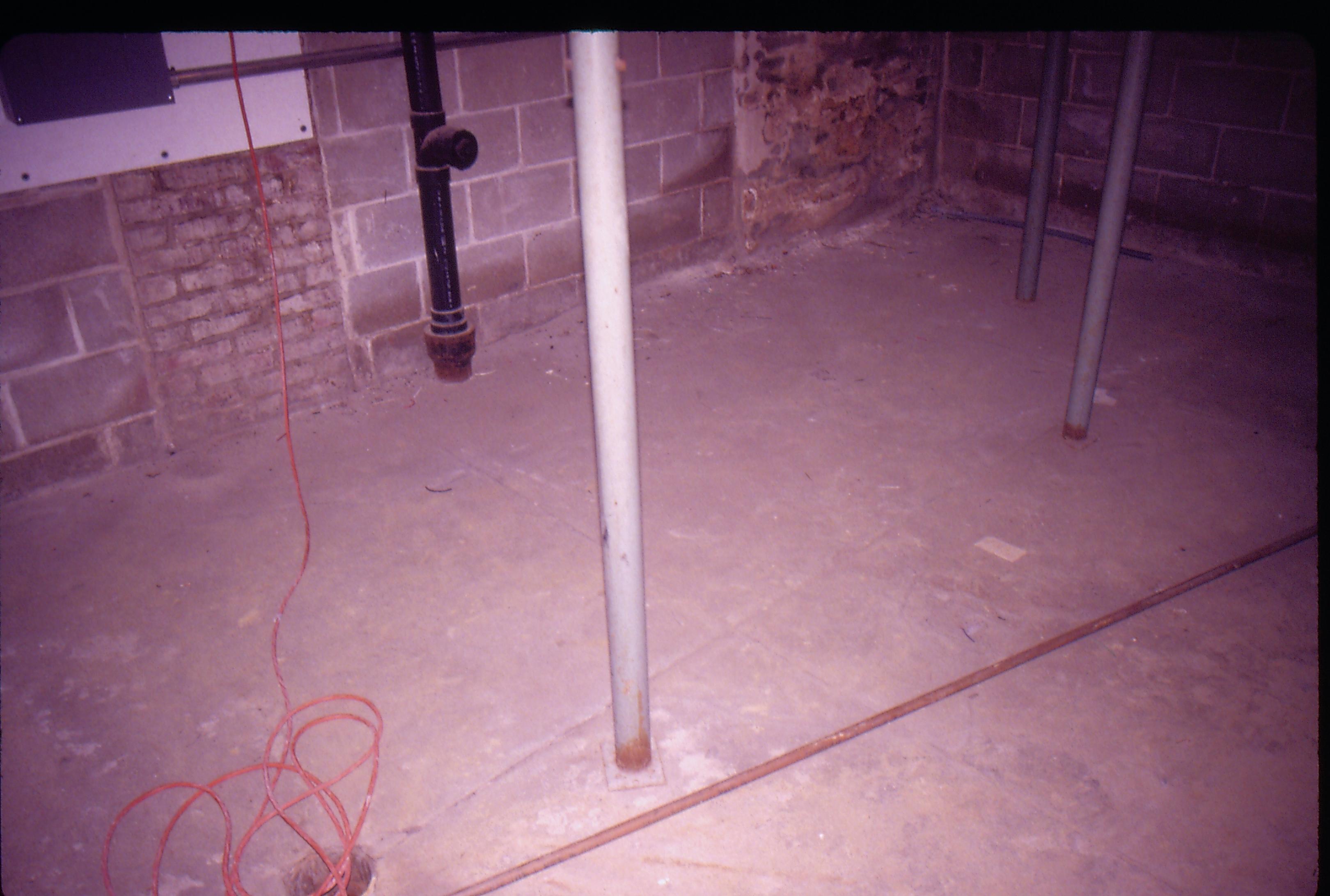 Lyon House - basement. Concrete floor with modern metal supports in center of room. Walls are a mix of old brick and modern cinder block.  Sewer pipe running down wall in background. Extension cords on left near Circuit Breaker box. This area is under kitchen on first floor Looking Northwest from basement Lyon, Basement, concrete, cinder block, breaker box, pipes