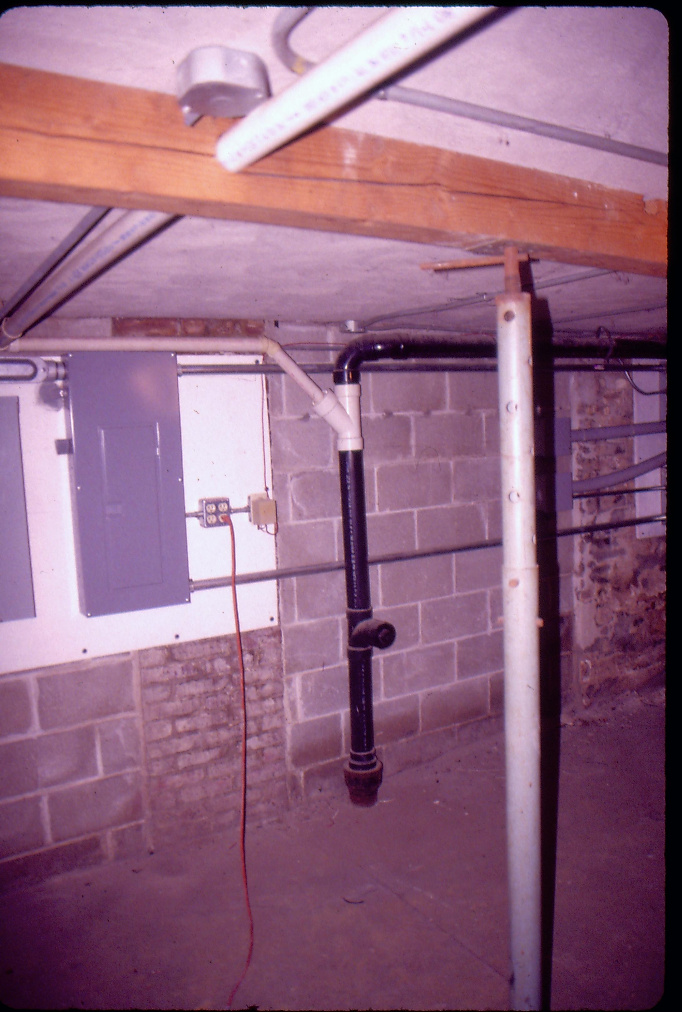 Lyon House - basement, PVC pipe in furnace room along back wall. Adjustable metal support in foreground right. Conduit, circuit break box and other utility equipment attached to back wall, a mix of old brick and modern cinder block. Floor is concrete. Wooden beam visible on ceiling with pipes and conduit running through it.  This area is under the kitchen on the 1st floor Looking Northwest from basement Lyon, Basement, utilities, PVC pipe, breaker box, cinder block, concrete