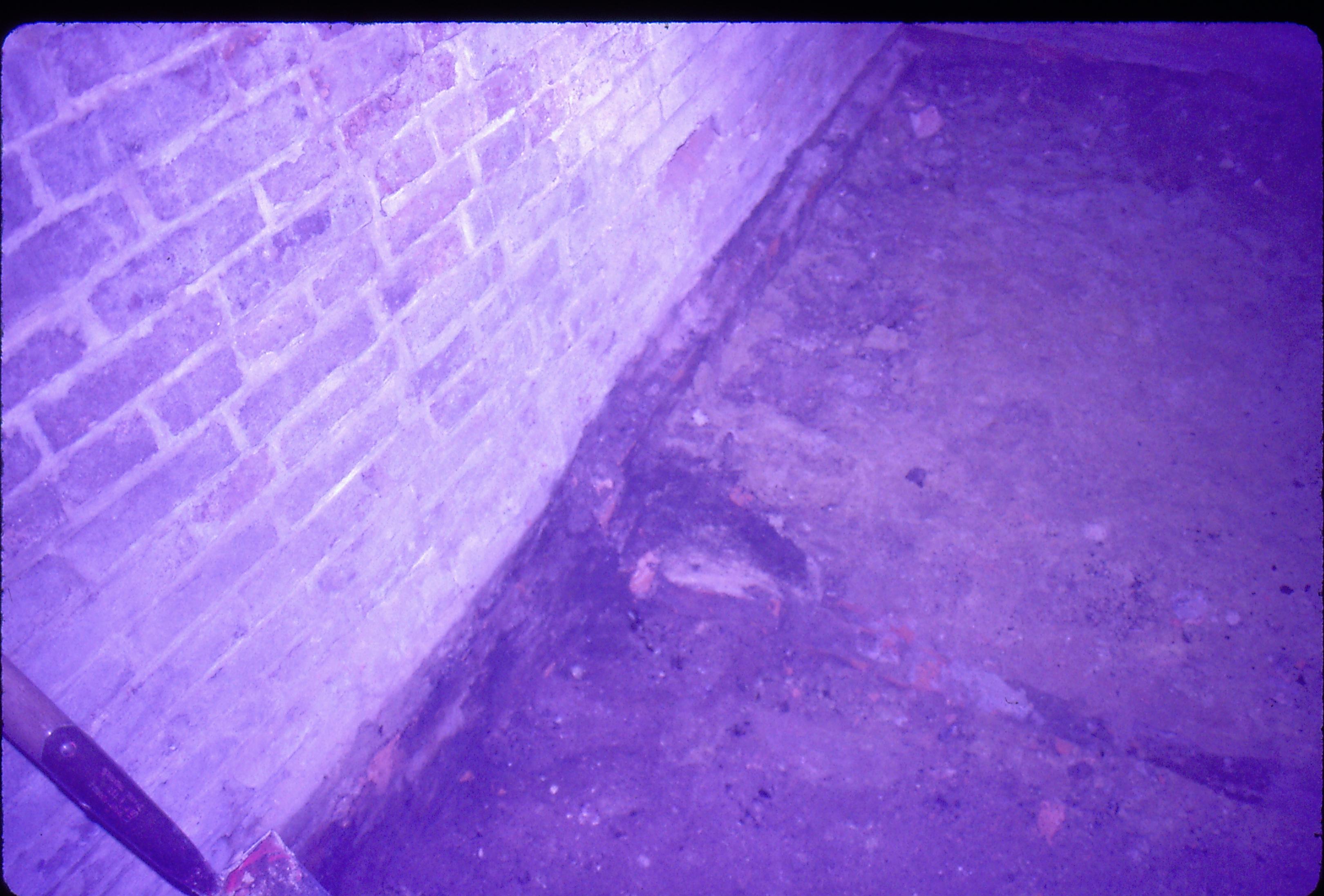 Lyon House - basement, closeup of wall where trench to cistern/gravity furnace? meets wall. Brick foot to brick foundation wall visible at joint between wall and dirt and brick rubble floor. Shovel leaning against wall in left foreground Looking East from basement Lyon, Basement, brick wall, foundation