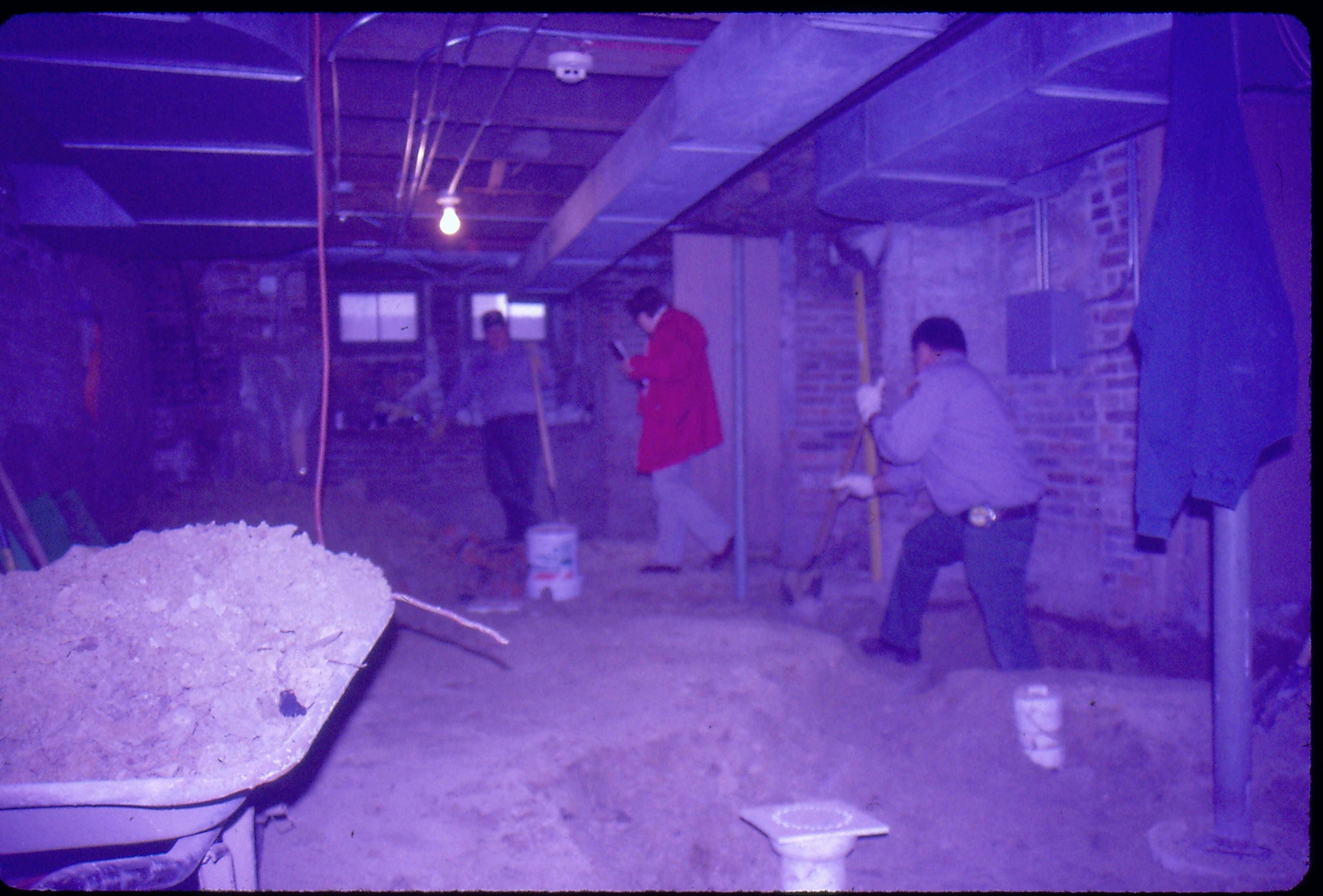 Lyon House - basement. Maintenance Workers Tom Melton? in background, and Tom Pacha dig out trench while Curator Susan Haake checks out the flooring near the doorway. Wheel barrow and square floor drain in foreground. Cords and other equipment scattered around. Circuit breaker box visible behind Tom Pacha Looking South/Southwest in basement Lyon, Basement, staff, duct work, trench, utilities