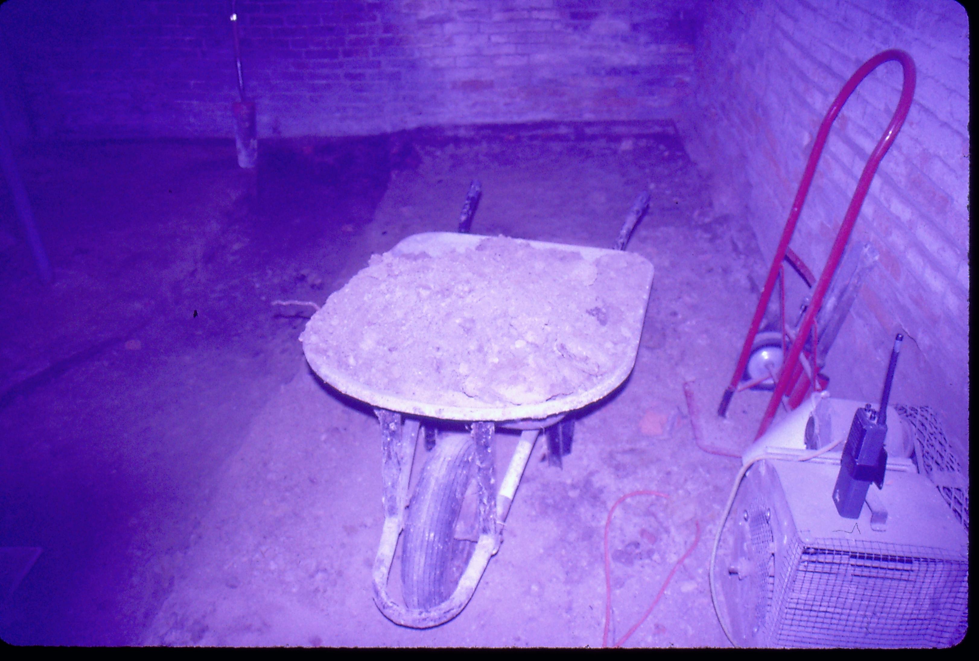 Lyon House - basement. Wheelbarrow filled with dirt and brick rubble from floor. Other equipment including dolly and park radio on right. Brick walls seen on right and background. Trench leading to cistern/gravity furnace? on left Looking North in basement Lyon, Basement, trench, equipment, brick walls