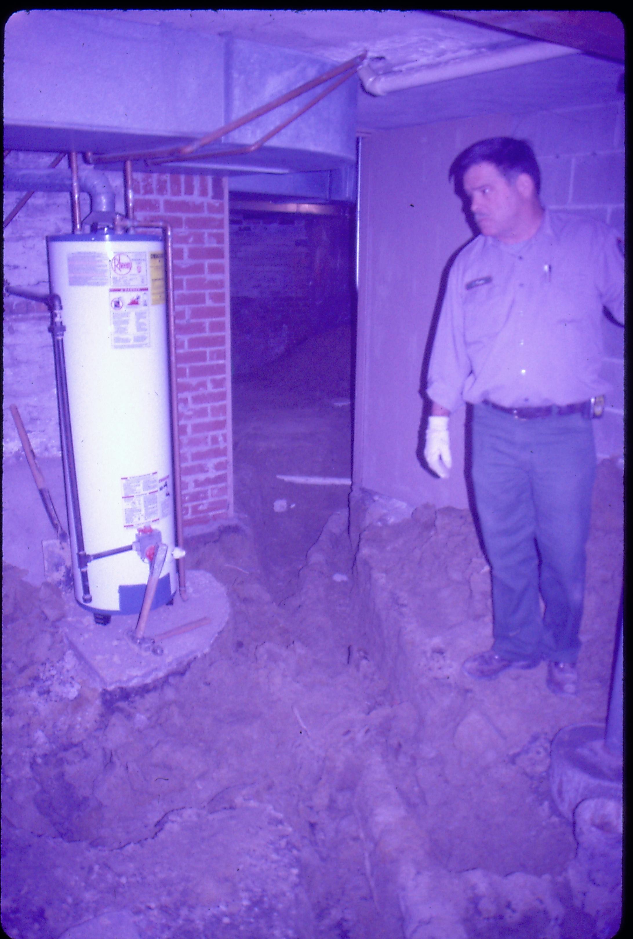 Lyon House - basement. Maintenance Worker Tom Pacha checks on water heater while digging up old plumbing pipes in dirt floor. Modern PVC pipe seen in background through doorway. Duct work on ceiling. Looking Southeast from furnace room in basement Lyon, Basement, water heater, staff, plumbing, cinder block, duct work