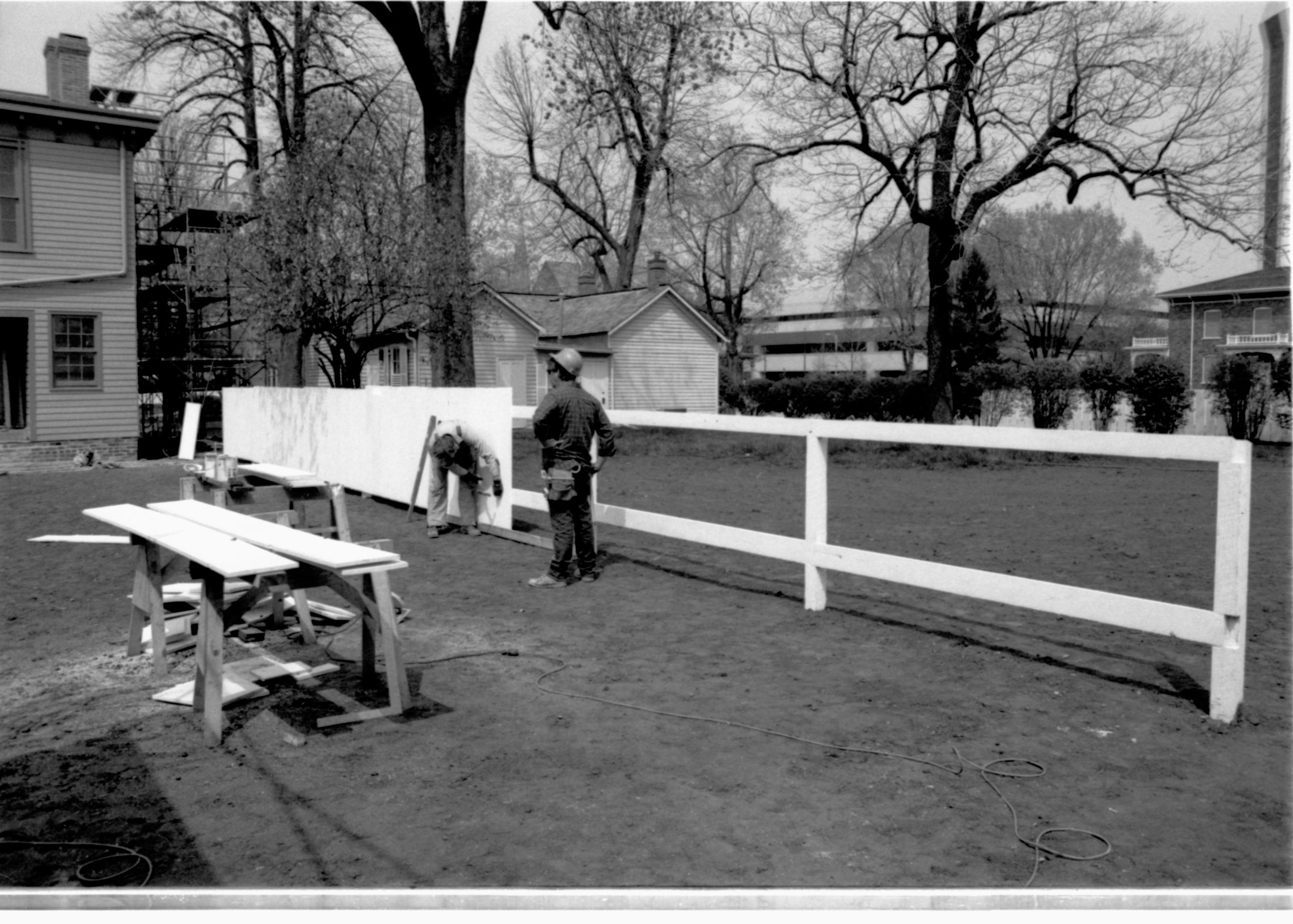 Two workers building fencing along the north line of the Lincoln Home lot. Lincoln Home in background, left; Corneau house (pre-move) in the background, center. Photographer facing northwest.
