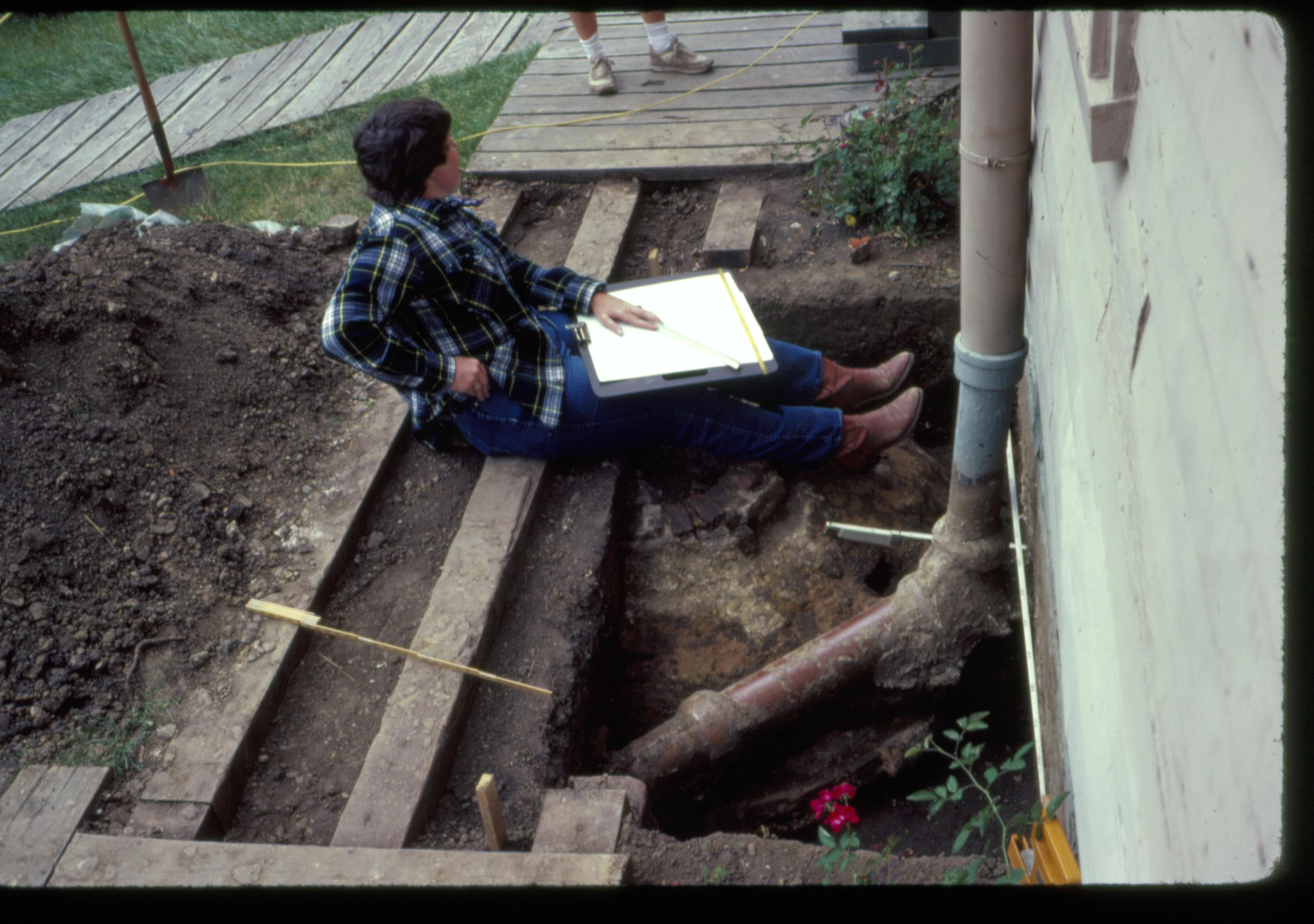 Cistern and water supply/roof drain of the Lincoln Home, in the back yard. Archeologist sits on boardwalk rails, speaking to a second person. Photographer facing south.