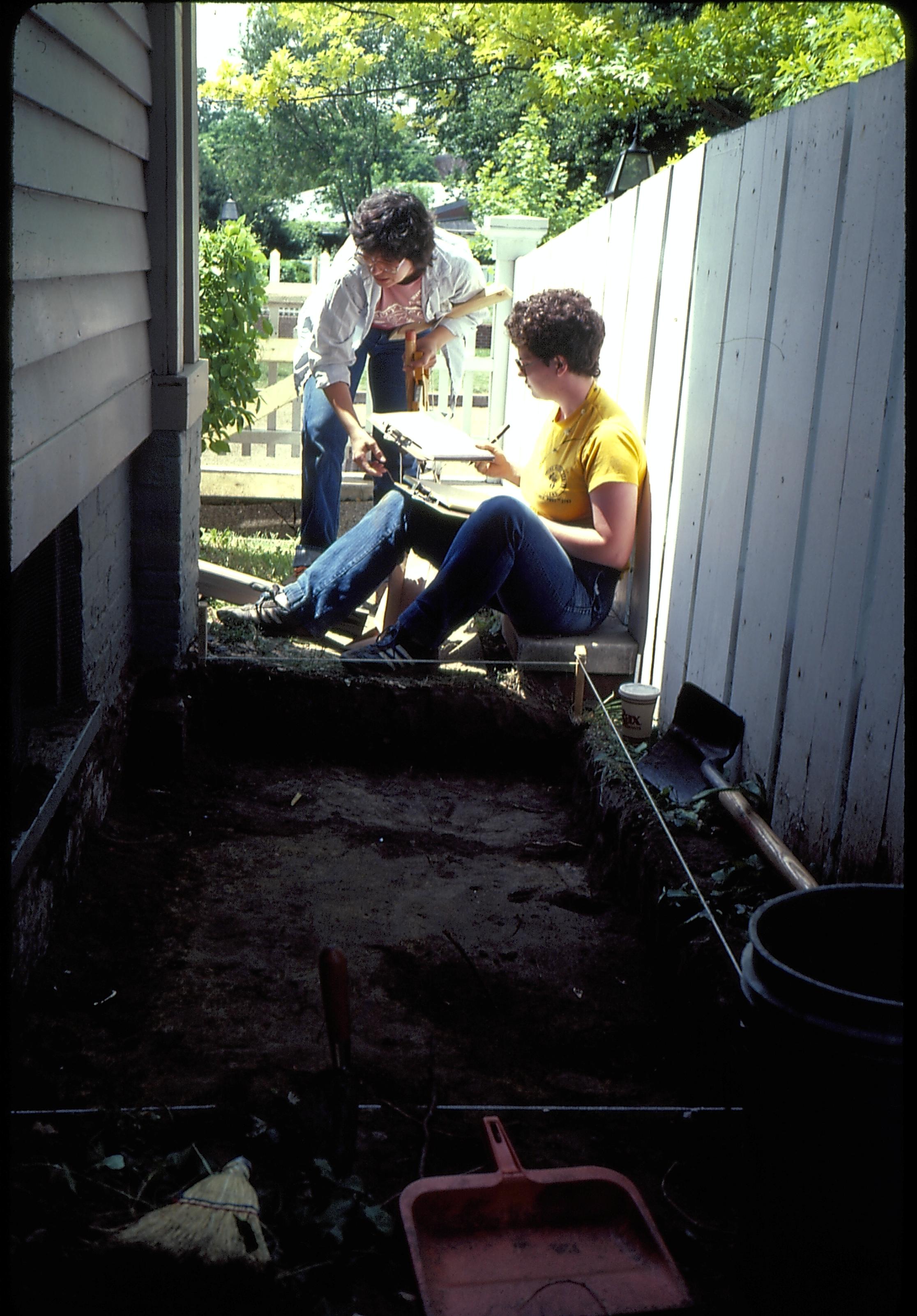 Archeology prior to the 1987 Lincoln Home Restoration. Two archeologists work on a test pit located along the north west corner of the Lincoln Home, north side. Photographer facing west.