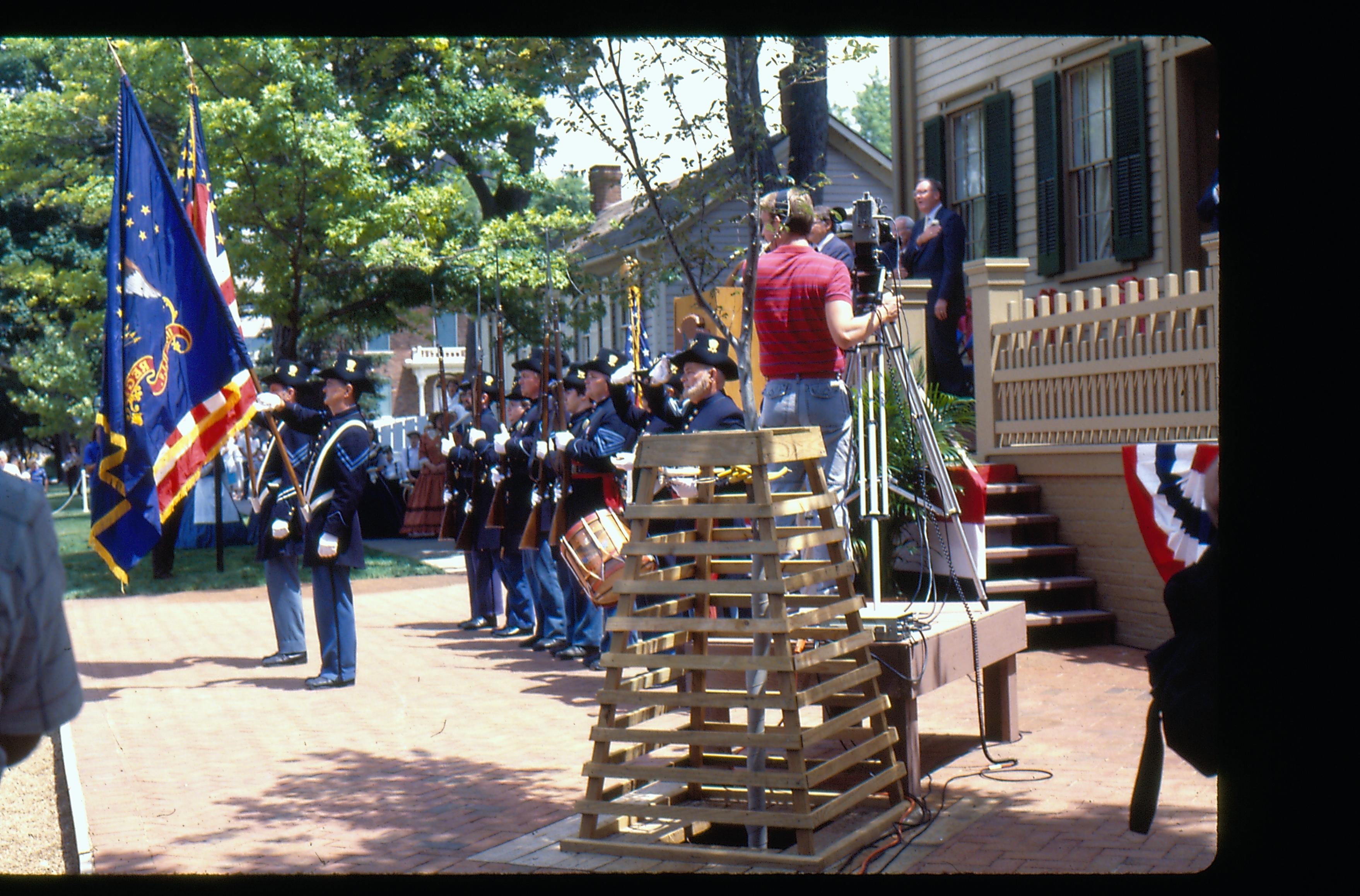 A Union Army reenactor group bearing US and regimental flags stand in formation in front of the Lincoln Home during reopening ceremony. A film crew can be seen recording the event in the middle foreground, along with a speakers podium. Dignitaries can be seen in the right background, standing in front of the restored Lincoln Home, at far right. The pre-move Corneau House can be seen in the background. Photographer facing north.