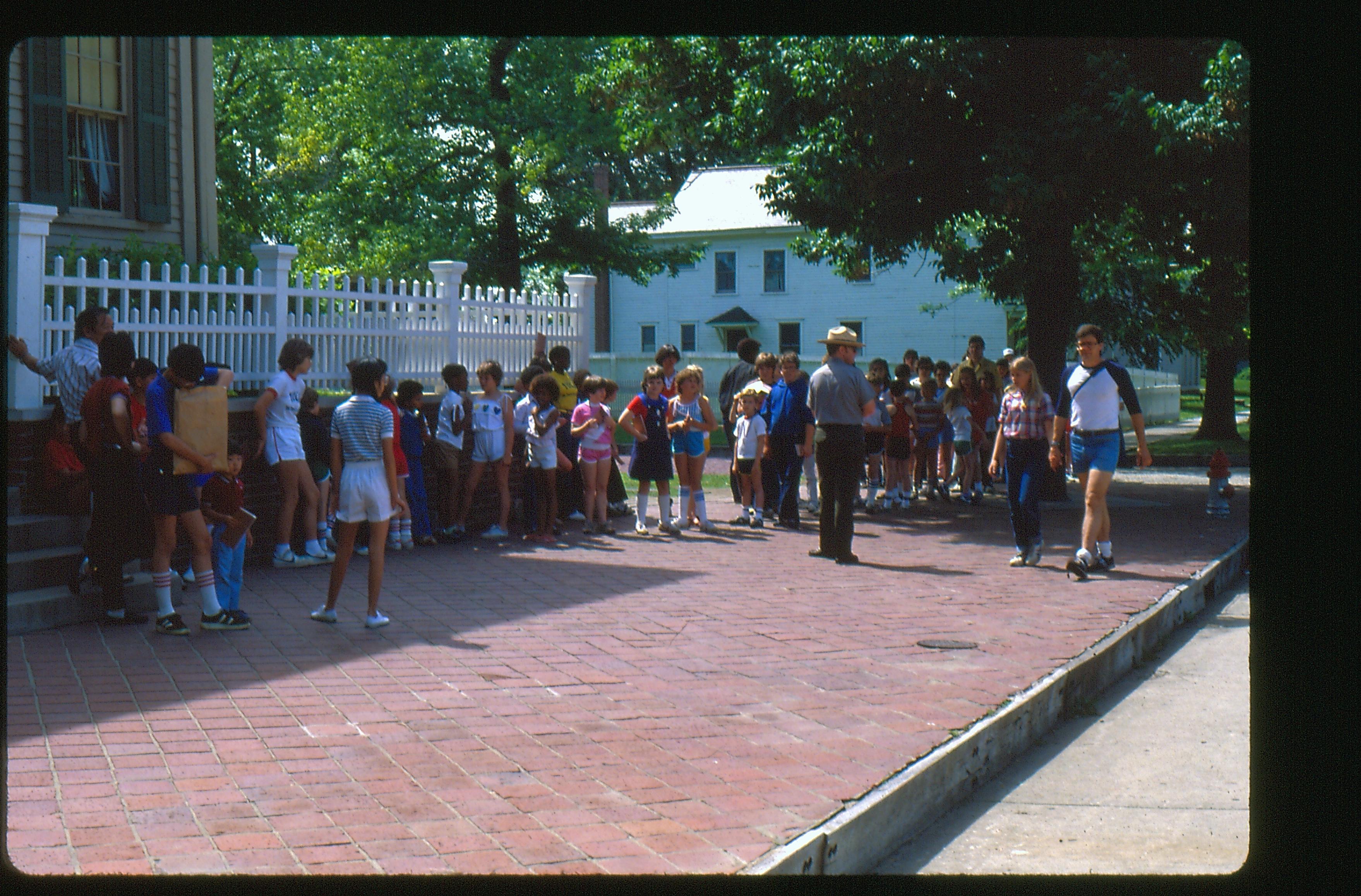 Visitors in a line heading south from the Lincoln Home. A single ranger waits alongside the group. Fire hydrant visible on brick plaza to far right of the photo. Cook house in background.