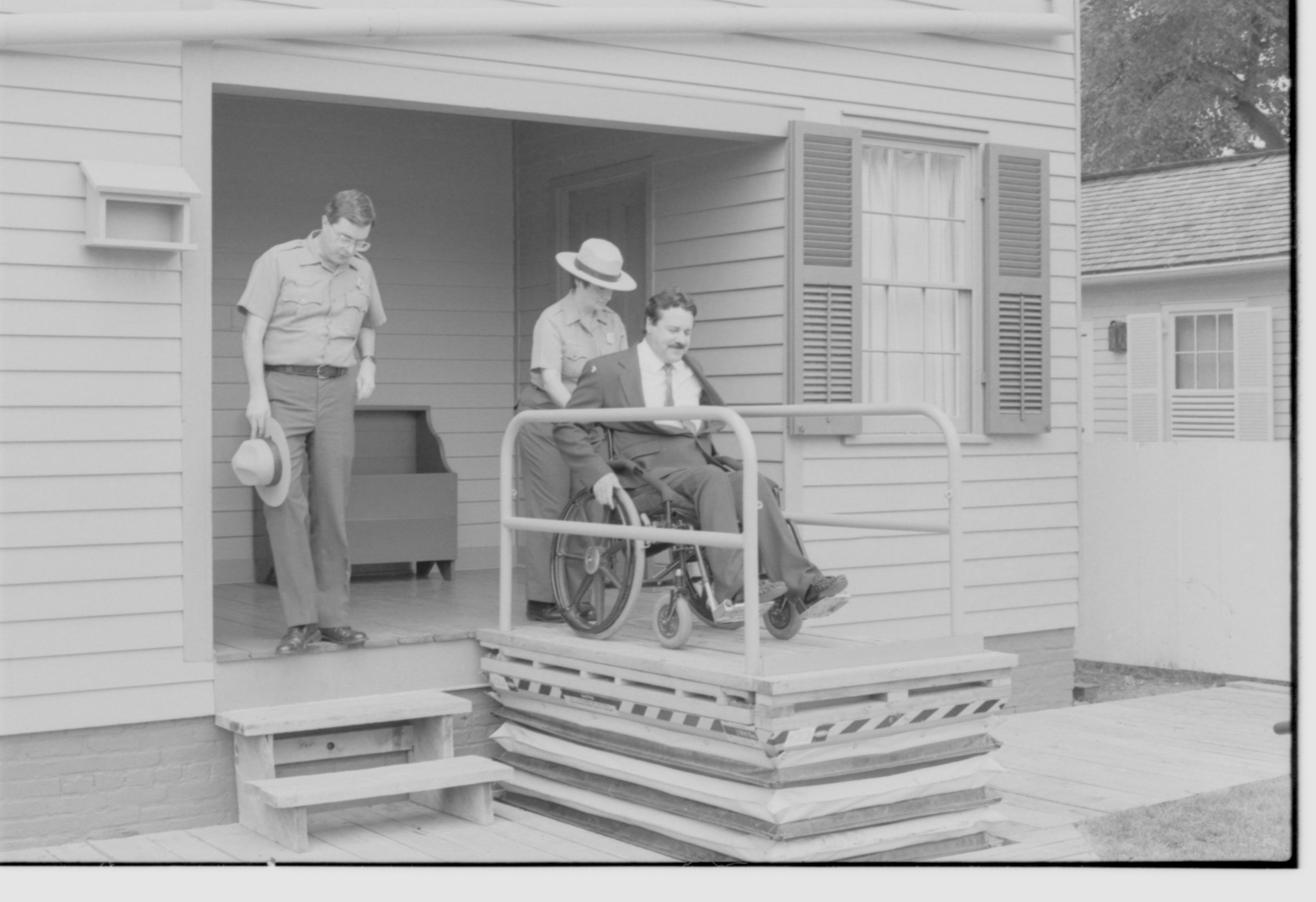 Lincoln Home wheelchair lift fully extended. One man in wheelchair entering lift. Staff member Judith Winkelmann on back porch assistimg wheelchair onto lift. Superintendent Norman Hellmers on left of photo, on porch. Corneau house in background. Photographer facing north west.