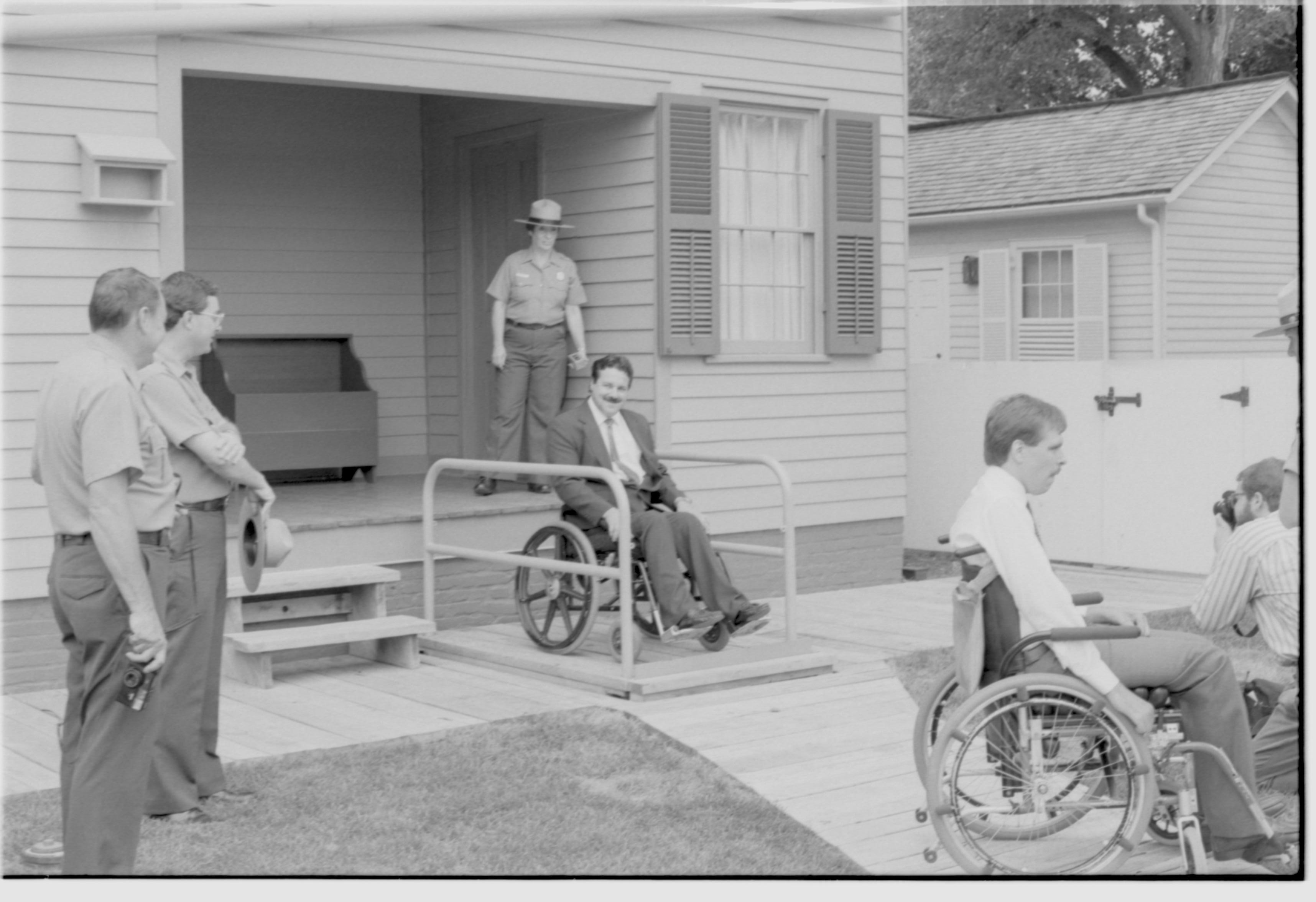 Lincoln Home wheelchair lift partially retracted. One man in wheelchair on lift, with one man in wheelchair and a photographer to right of picture. Staff member Judith Winkelmann on back porch using lift controls. Superintendent Norman Hellmers and second staff member on left of photo. Corneau house in background. Photographer facing north west.