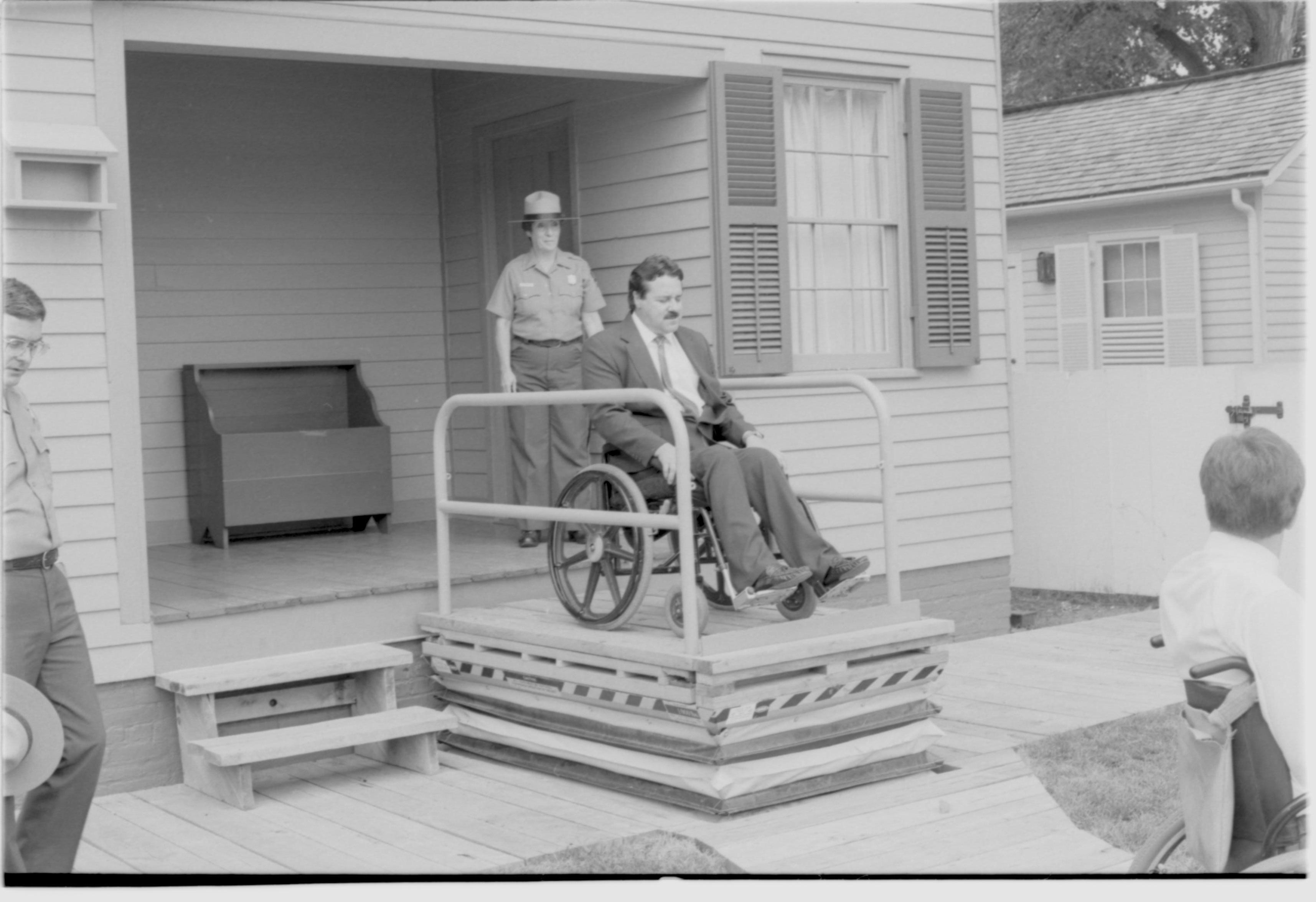 Lincoln Home wheelchair lift partially extended. One man in wheelchair on lift, with one man in wheelchair to right of picture. Staff member Judith Winkelmann on back porch using lift controls. Superintendent Norman Hellmers on left of photo. Corneau house in background. Photographer facing north west.