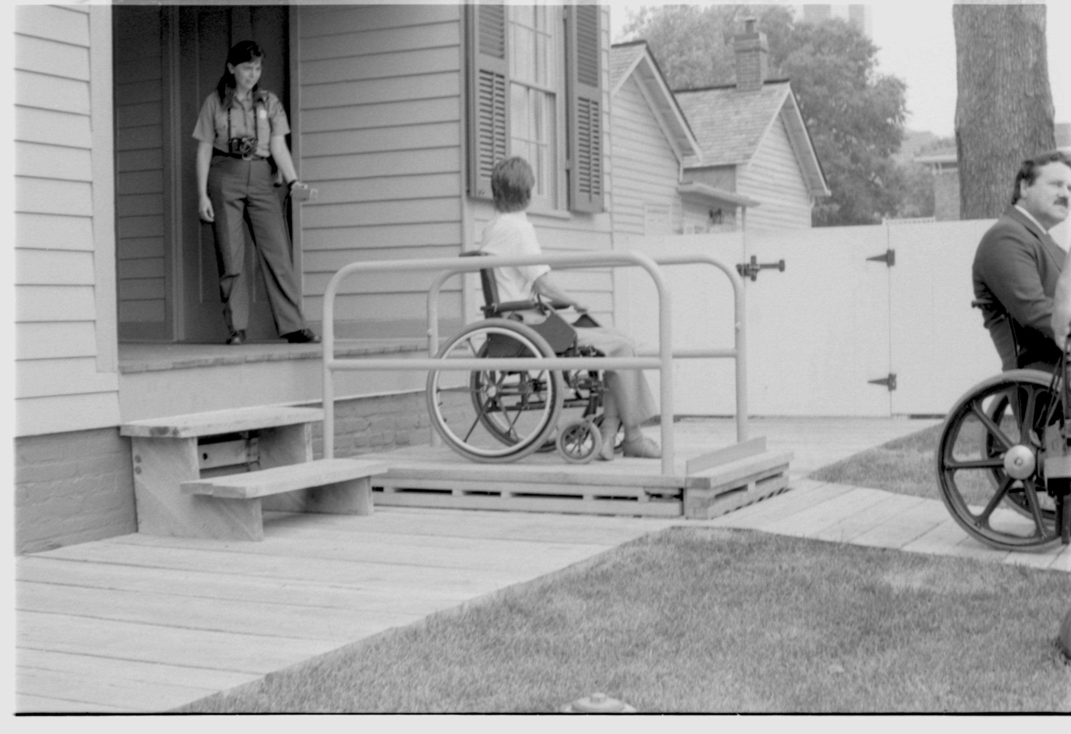 Lincoln Home wheelchair lift patially retracted. One man in wheelchair on lift, with one other man in wheelchair to right of picture. Staff member on back porch using lift controls. Corneau house in background. Photographer facing north west.