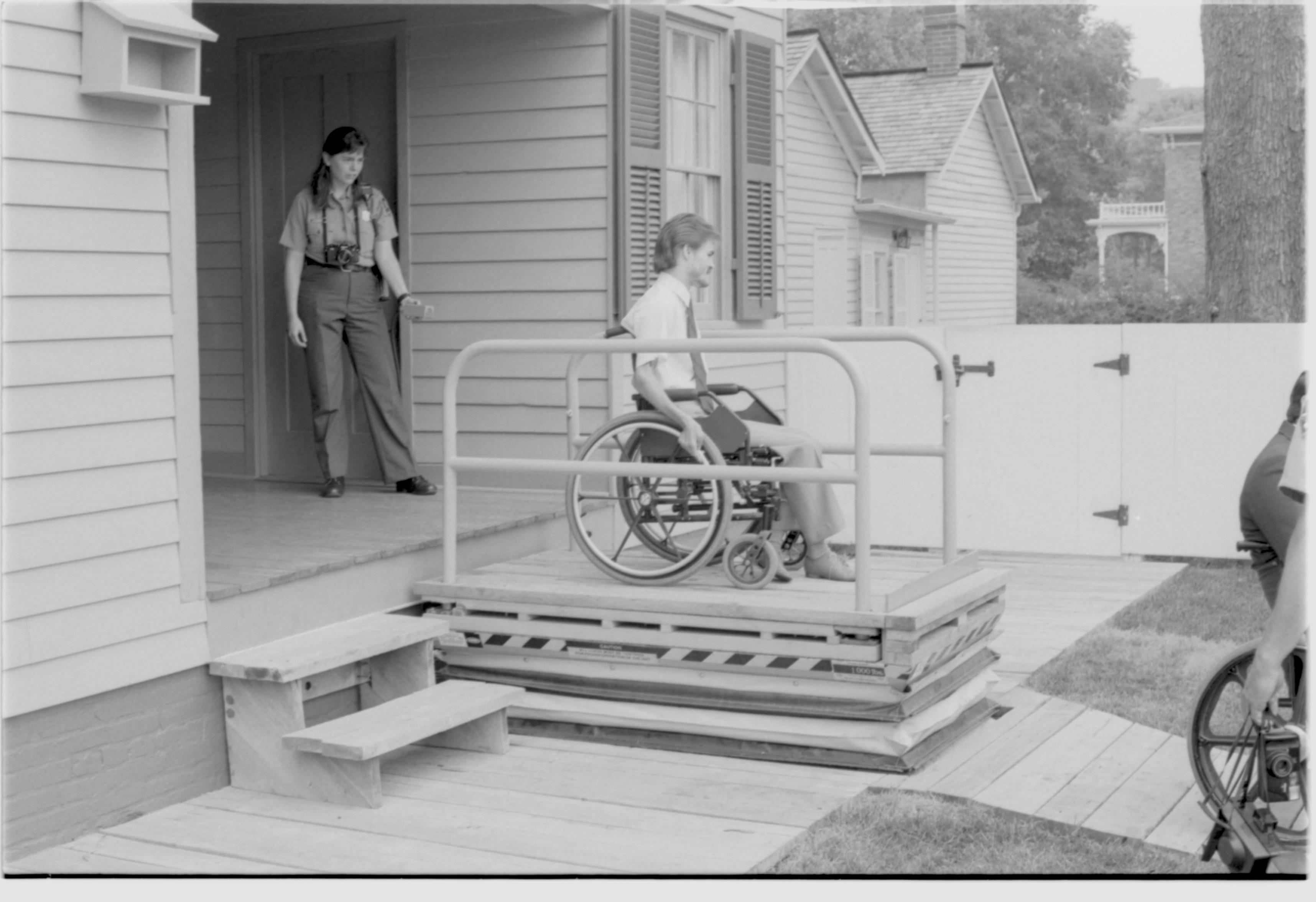 Lincoln Home wheelchair lift partially extended. One man in wheelchair on lift, with one other man in wheelchair to right of picture. Staff member on back porch using lift controls. Corneau house in background. Photographer facing north west.