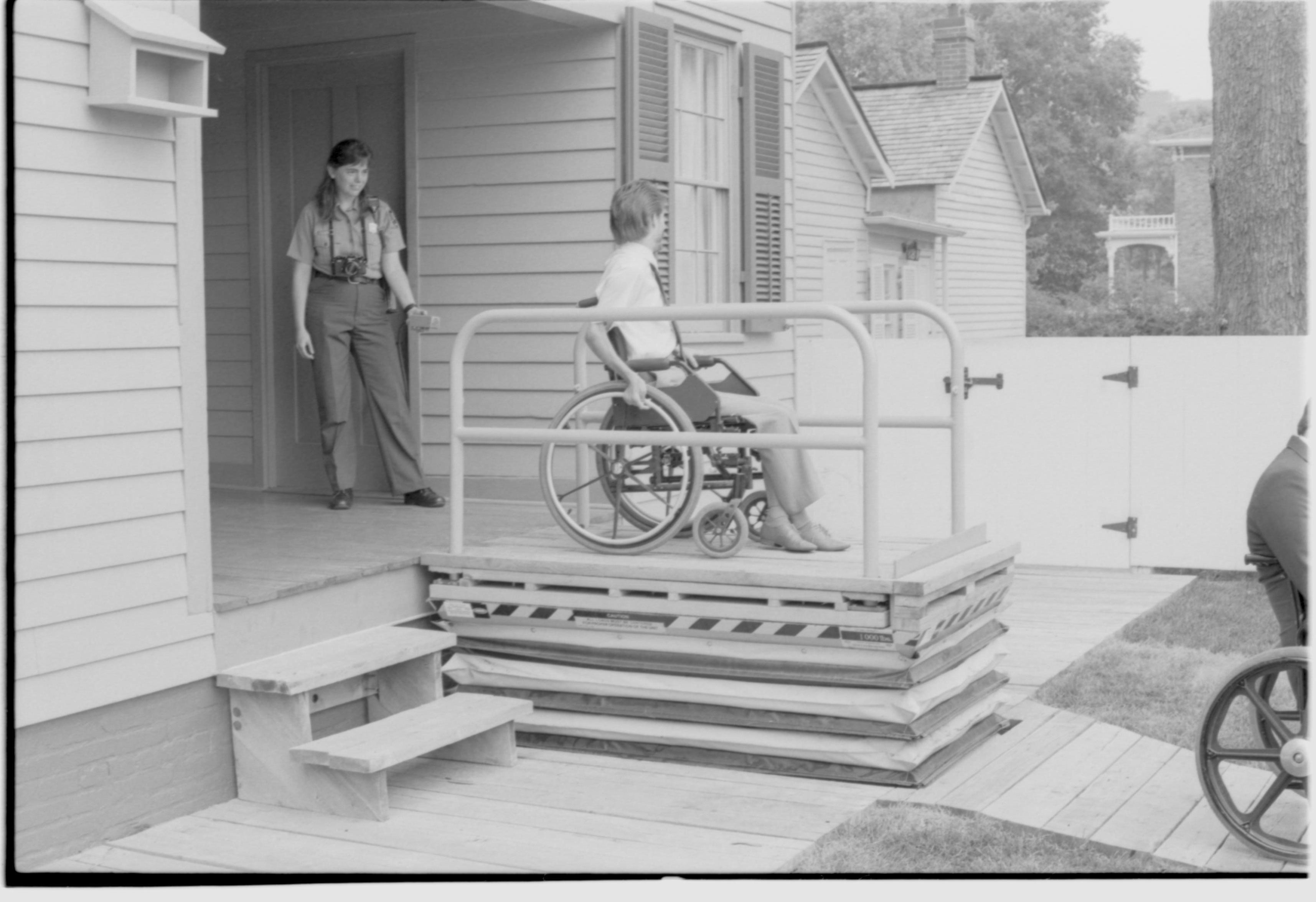 Lincoln Home wheelchair lift fully extended. One man in wheelchair on lift, with one other man in wheelchair to right of picture. Staff member on back porch using lift controls. Corneau house in background. Photographer facing north west.