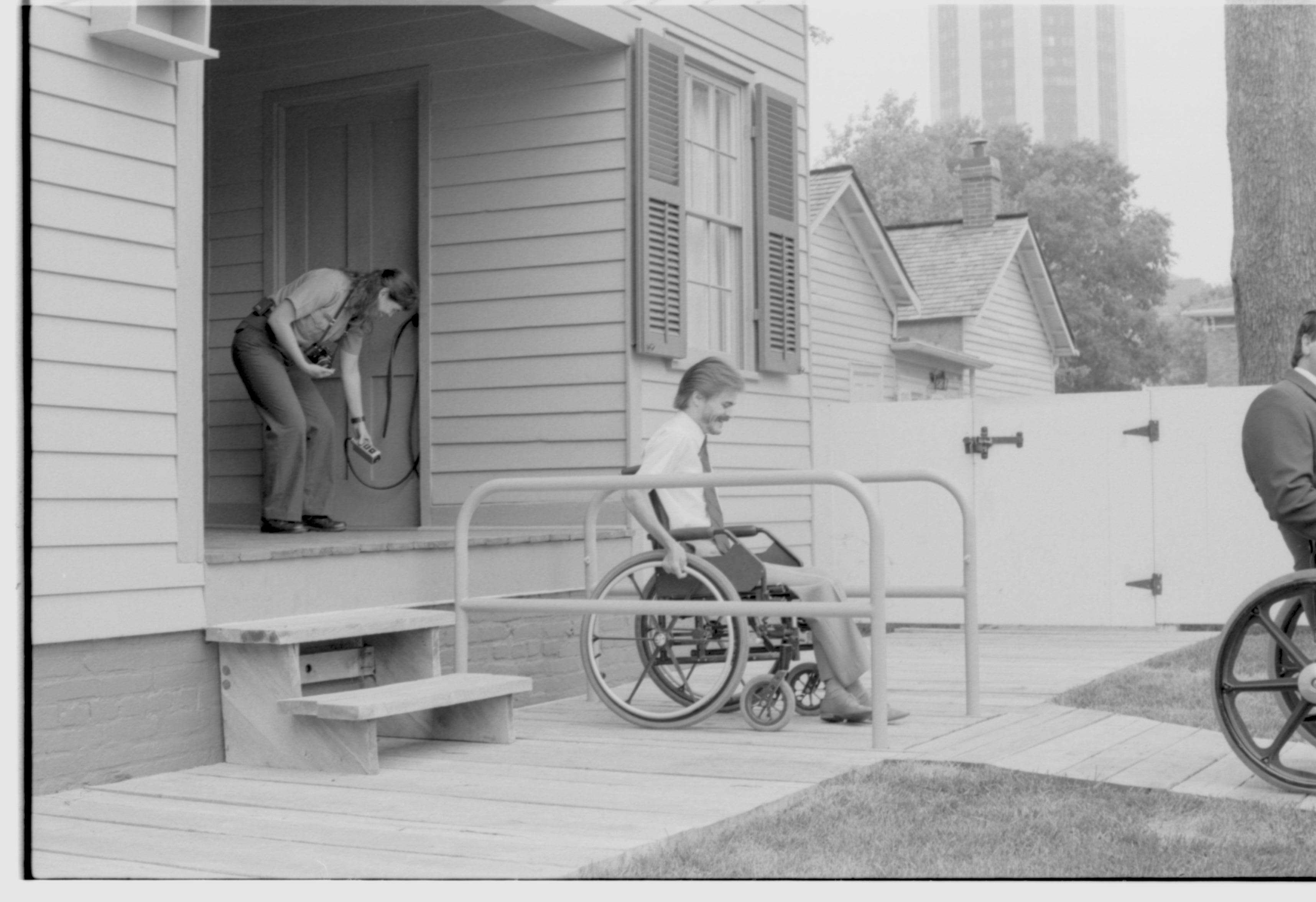 Lincoln Home wheelchair lift fully retracted. One man in wheelchair preparing to exit lift, with one other man in wheelchair to right of picture. Staff member on back porch using lift controls. Corneau house in background. Photographer facing north west.