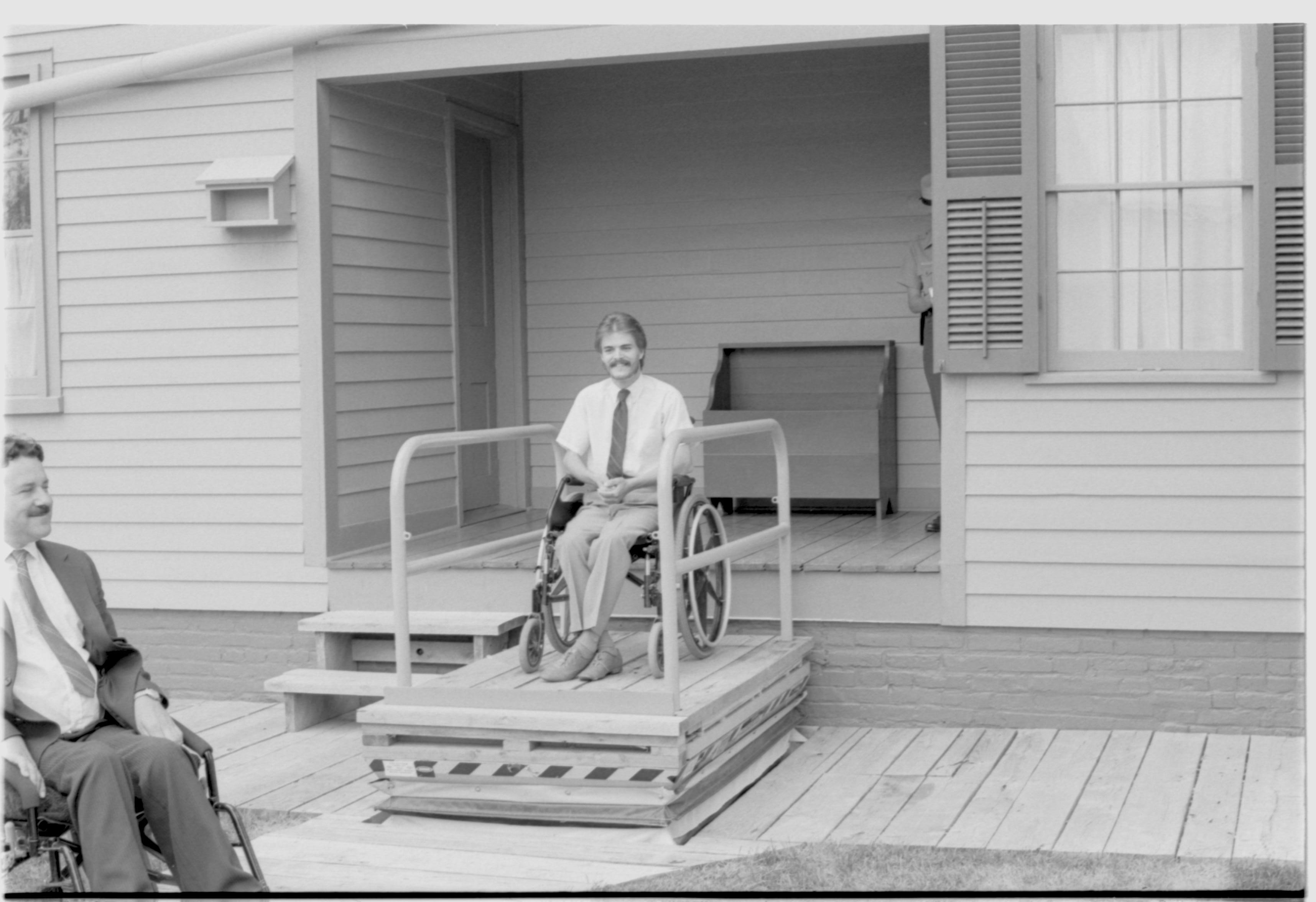 Lincoln Home wheelchair lift, at half height. One man in wheelchair on lift, facing away from Lincoln Home, with second man in wheelchair at ground level. Photographer facing south west.