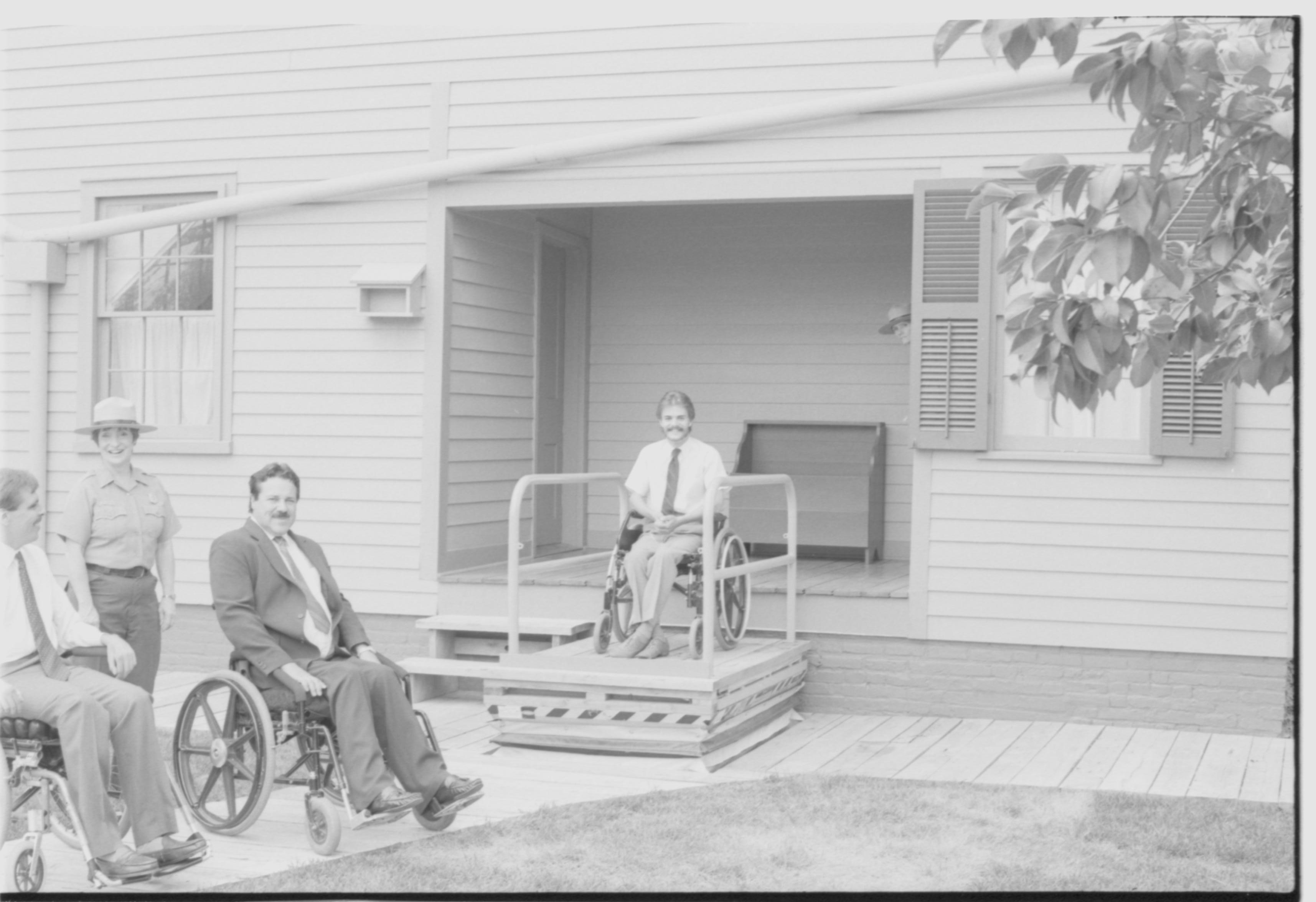 Lincoln Home wheelchair lift, at half height. One man in wheelchair on lift, facing away from Lincoln Home, with two other men in wheelchairs at ground level. Staff member Judith Winkelmann stands behind men in wheelchairs at ground level. Photographer facing south west.