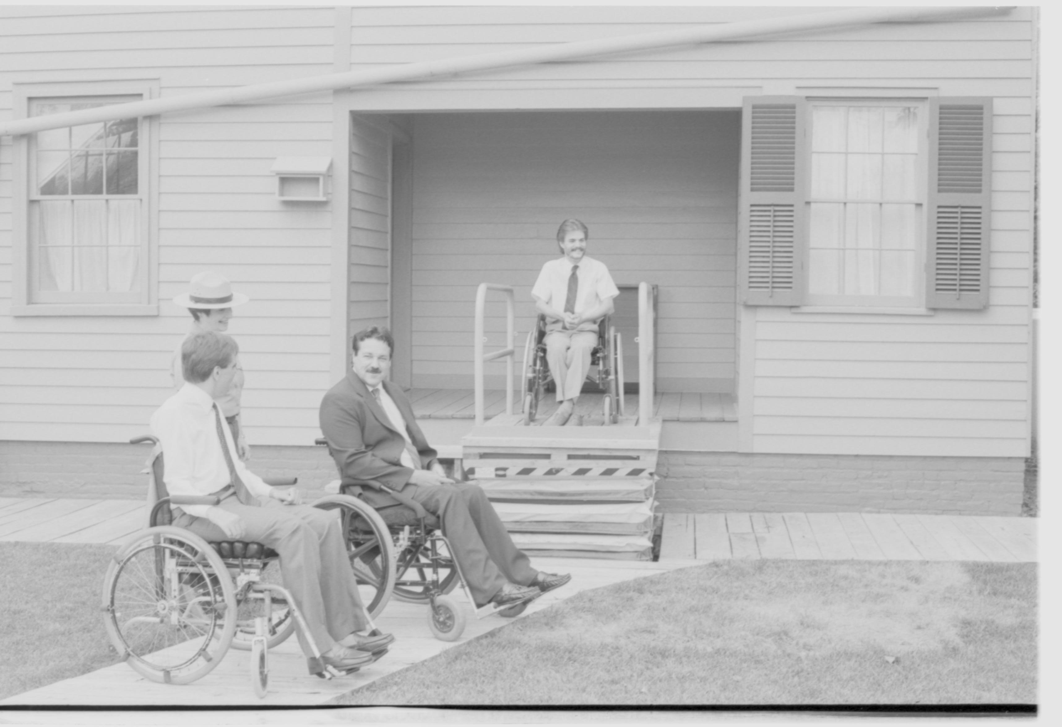 Lincoln Home wheelchair lift, at full height. One man in wheelchair on lift, facing away from Lincoln Home, with two other men in wheelchairs at ground level. Staff member Judith Winkelmann stands behind men at ground level. Photographer facing west.