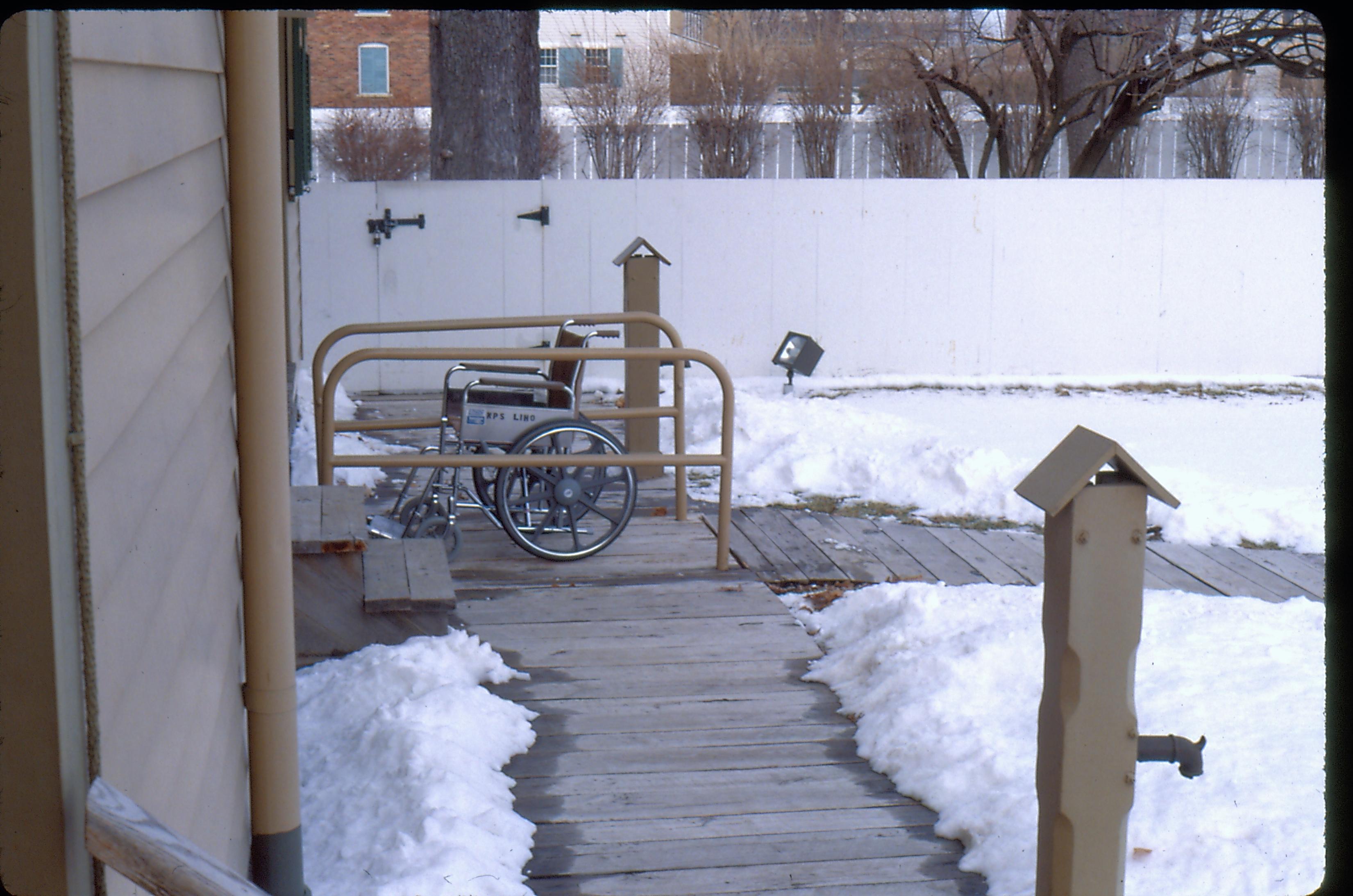 View of the Lincoln Home wheelchair lift, with empty wheelchair and rails. Snow on the ground. Photographer facing north.