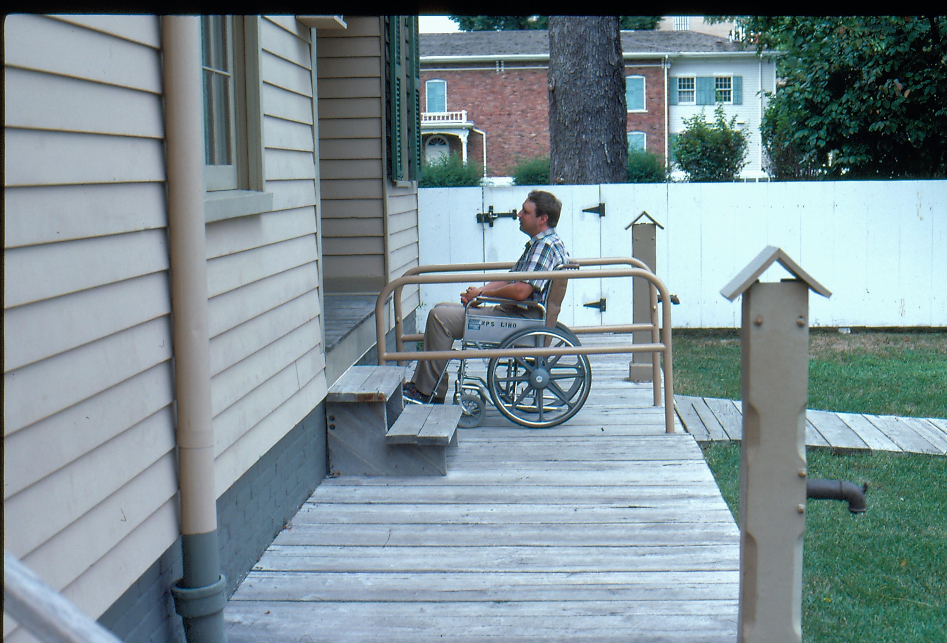 View of the Lincoln Home wheelchair lift, fully retracted without rails. Visitor on wheel chair present on lift, facing west. Photographer facing north.