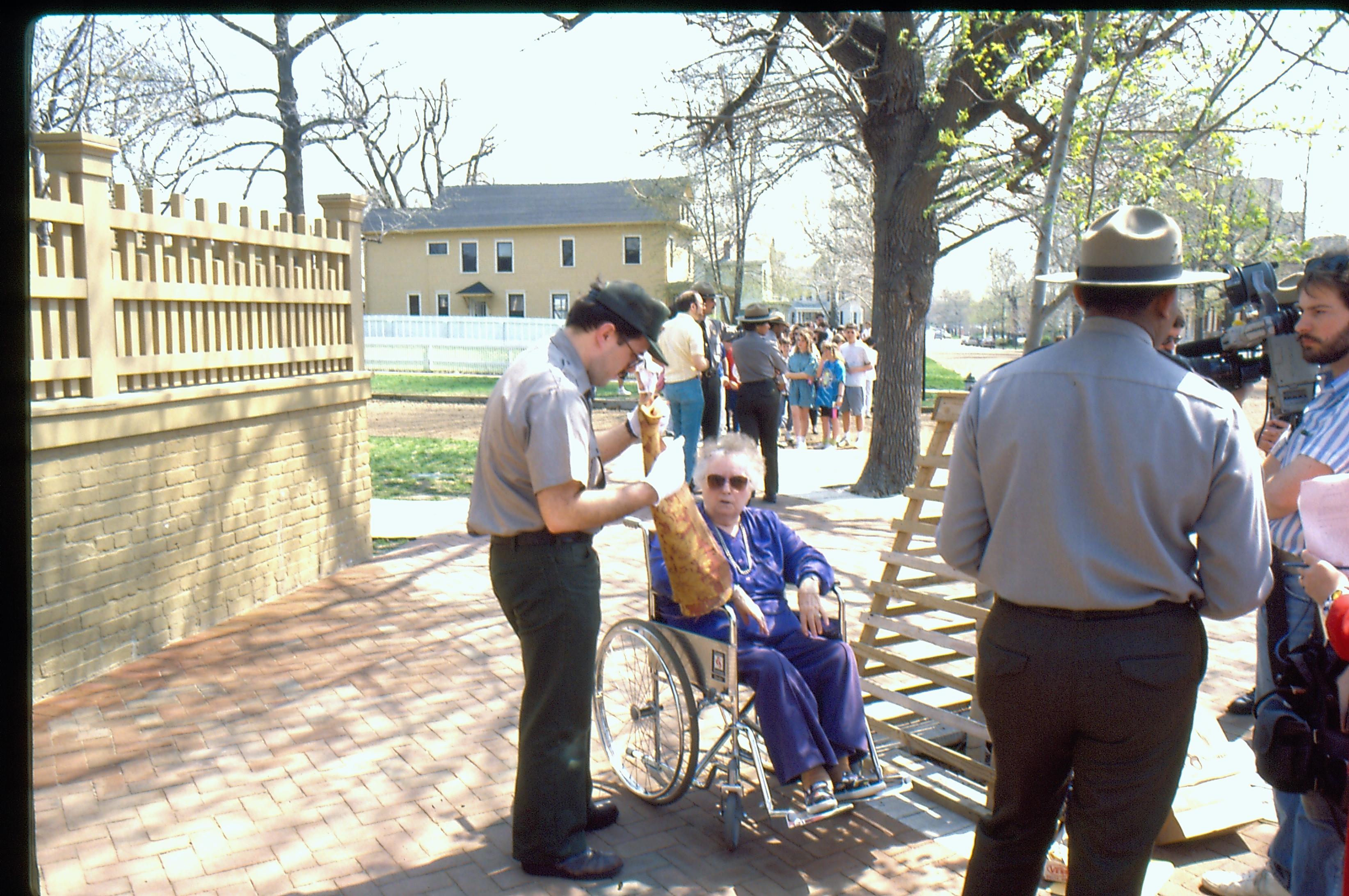 Two rangers assist a visitor in a wheelchair, as a news crew looks on. Third ranger with group in the background. Cook house in background, center. Photographer facing south east.