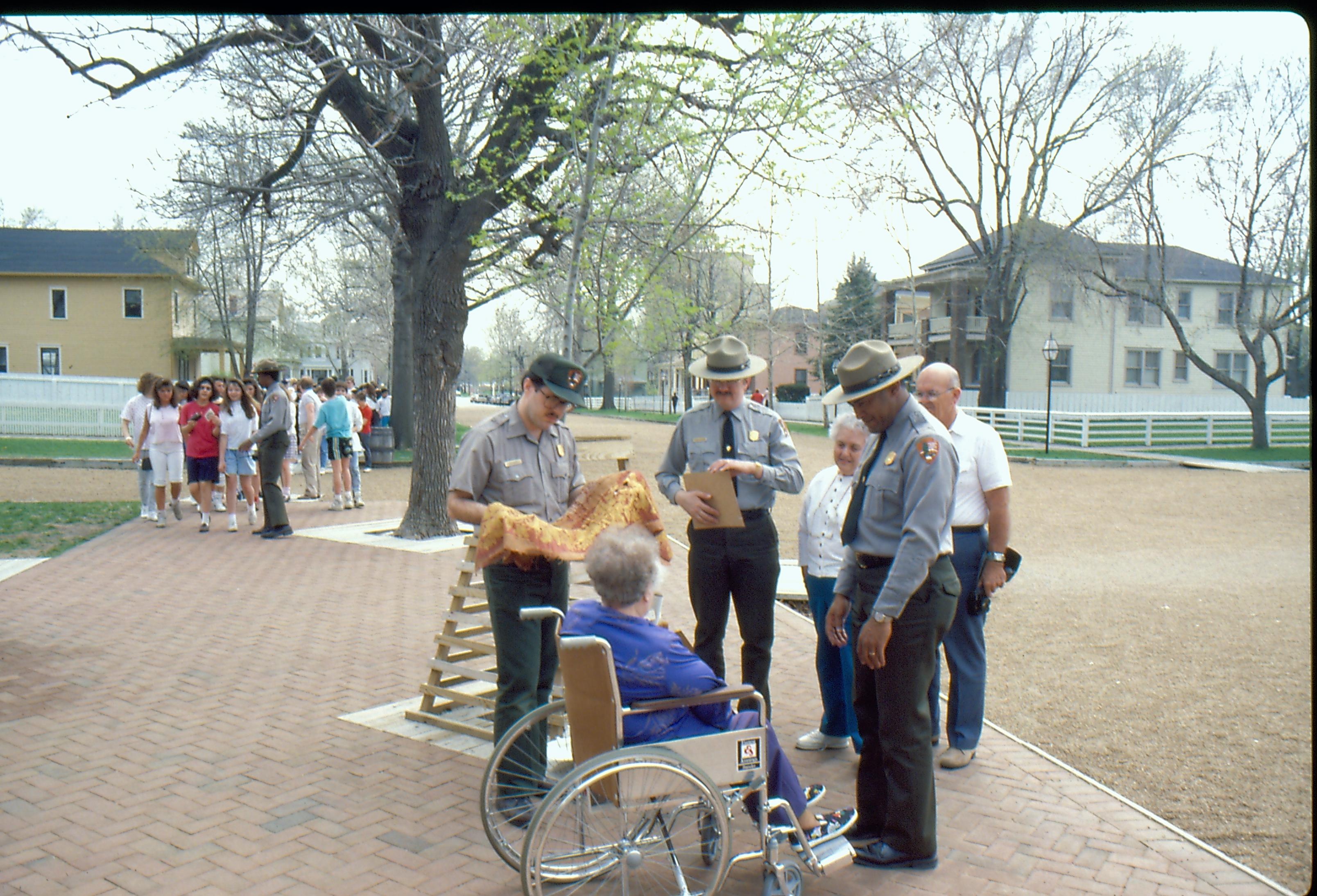 Three rangers, with fourth in background, assist a visitor using a wheelchair in front of the Lincoln Home. Cook house in background left, with pre-restoration Sprigg in background, right. Photographer facing south.
