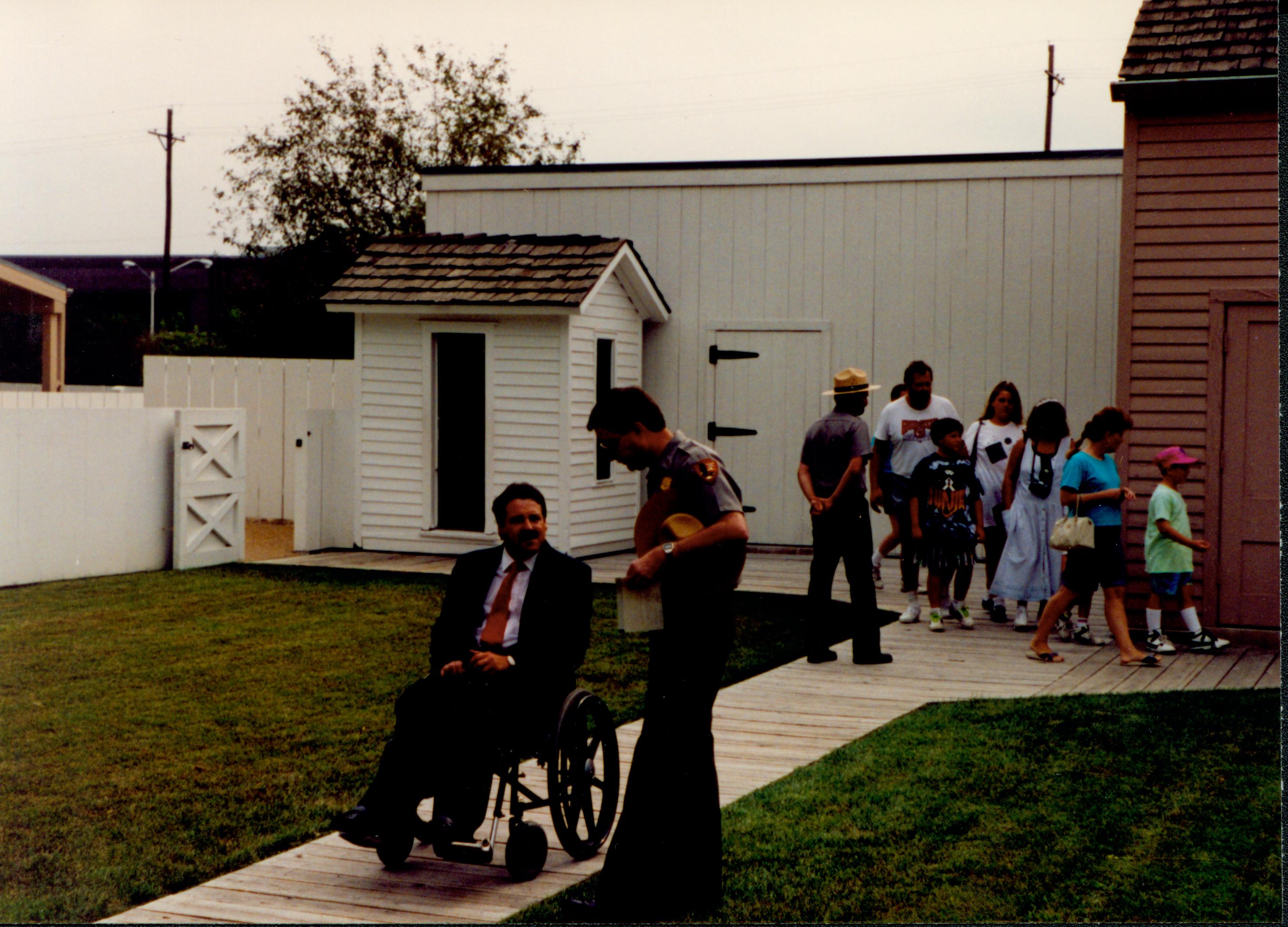 A man in a wheelchair waits on the Lincoln home back yard boardwalk while chatting with a Lincoln Home staff member. Visitors in the background. Photographer facing north east.