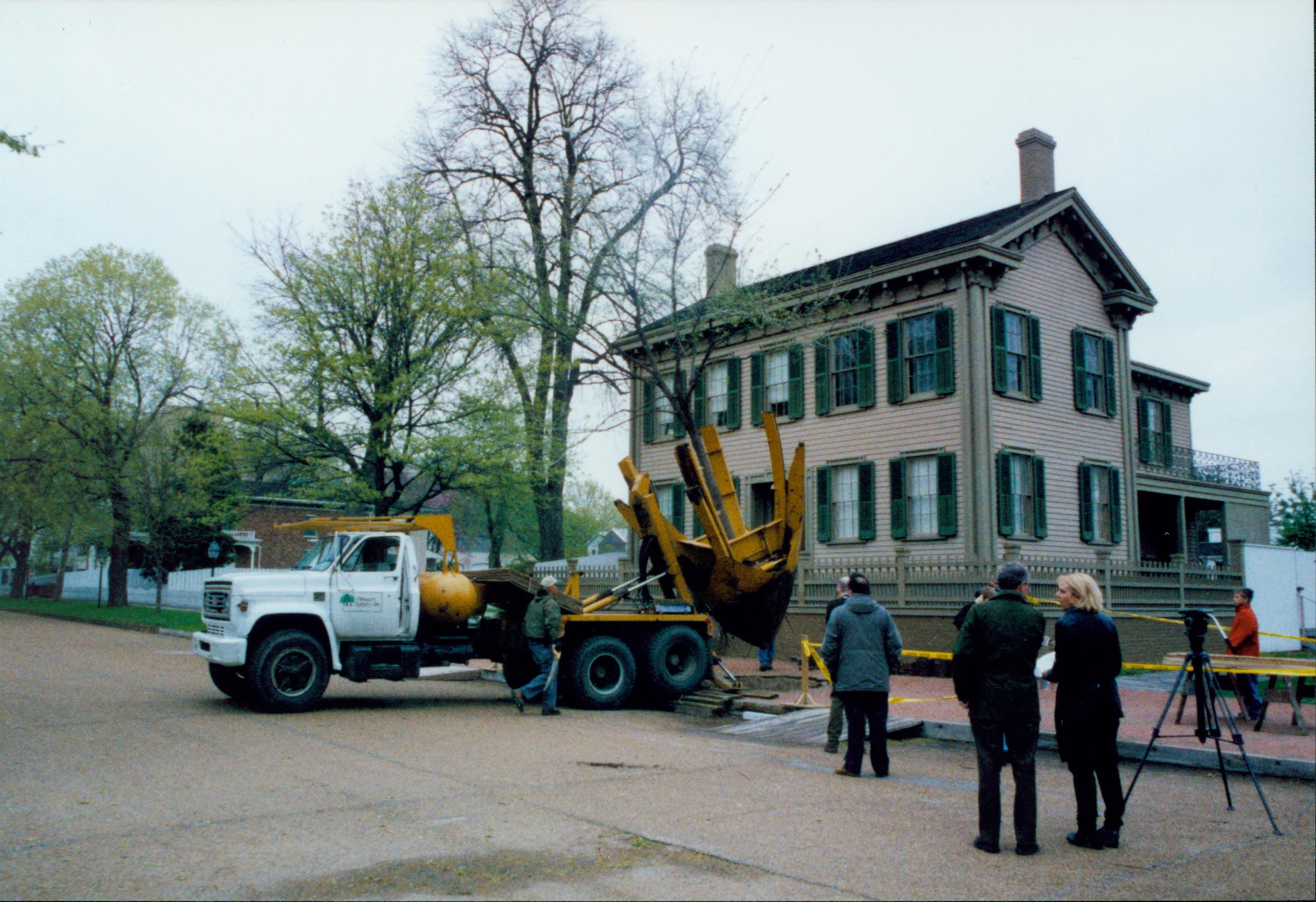 Elm tree replacement - removing tree from brick plaza in front of Lincoln Home. Superintendent Norman Hellmers speaks to reporter on right.  Pleasant Nursery staff and visitors watch. Conference Center in background far left Looking Northeast from 8th and Jackson Street intersection Elm tree, Lincoln Home, Conference Center, contractors, staff, reporters, brick plaza, Pleasant Nursery