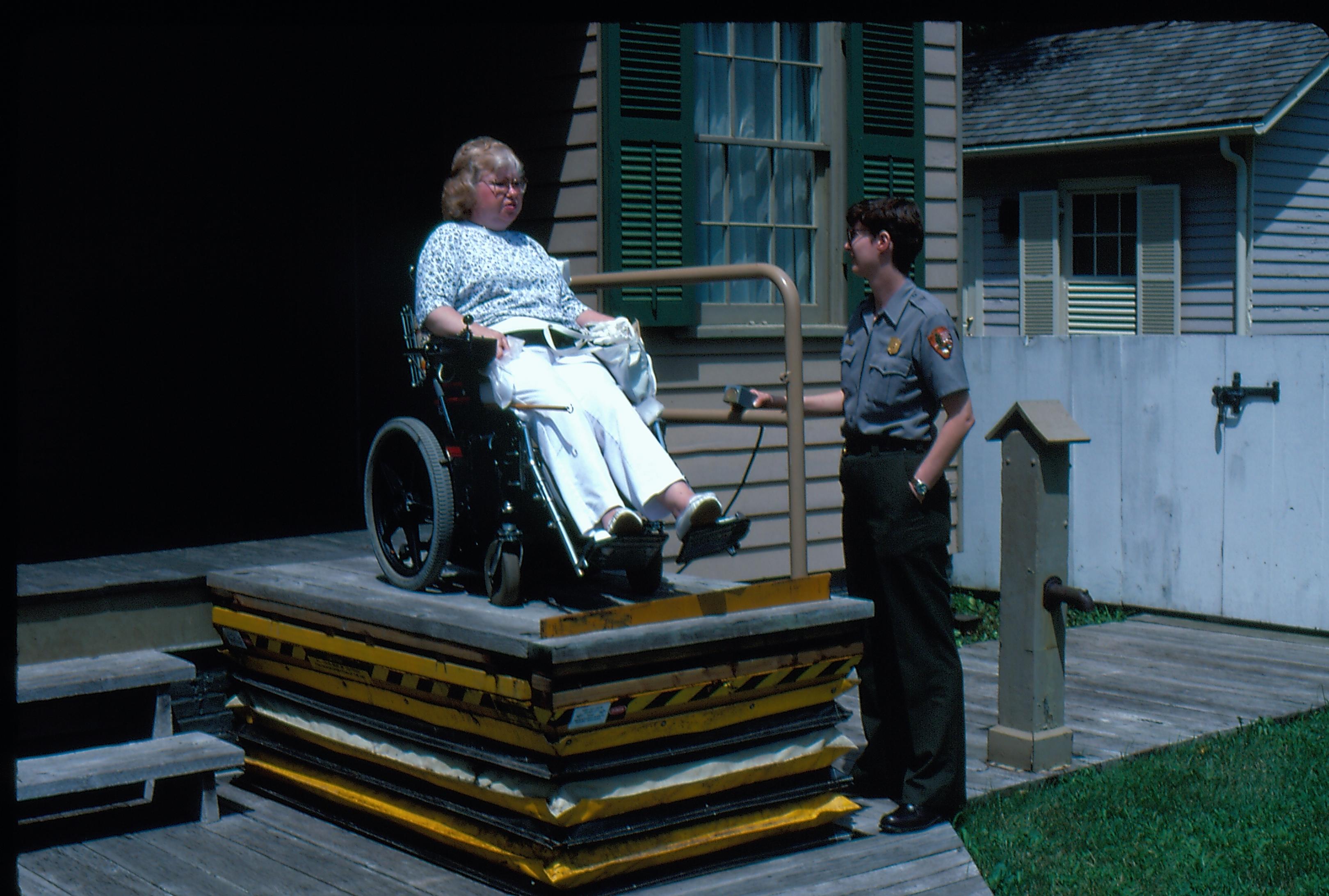 Lincoln Home wheelchair lift fully extended, with one handrail missing. One woman in wheelchair on lift. Staff member Cathy Mancuso at ground level using lift controls. Corneau house in background. Photographer facing north west
