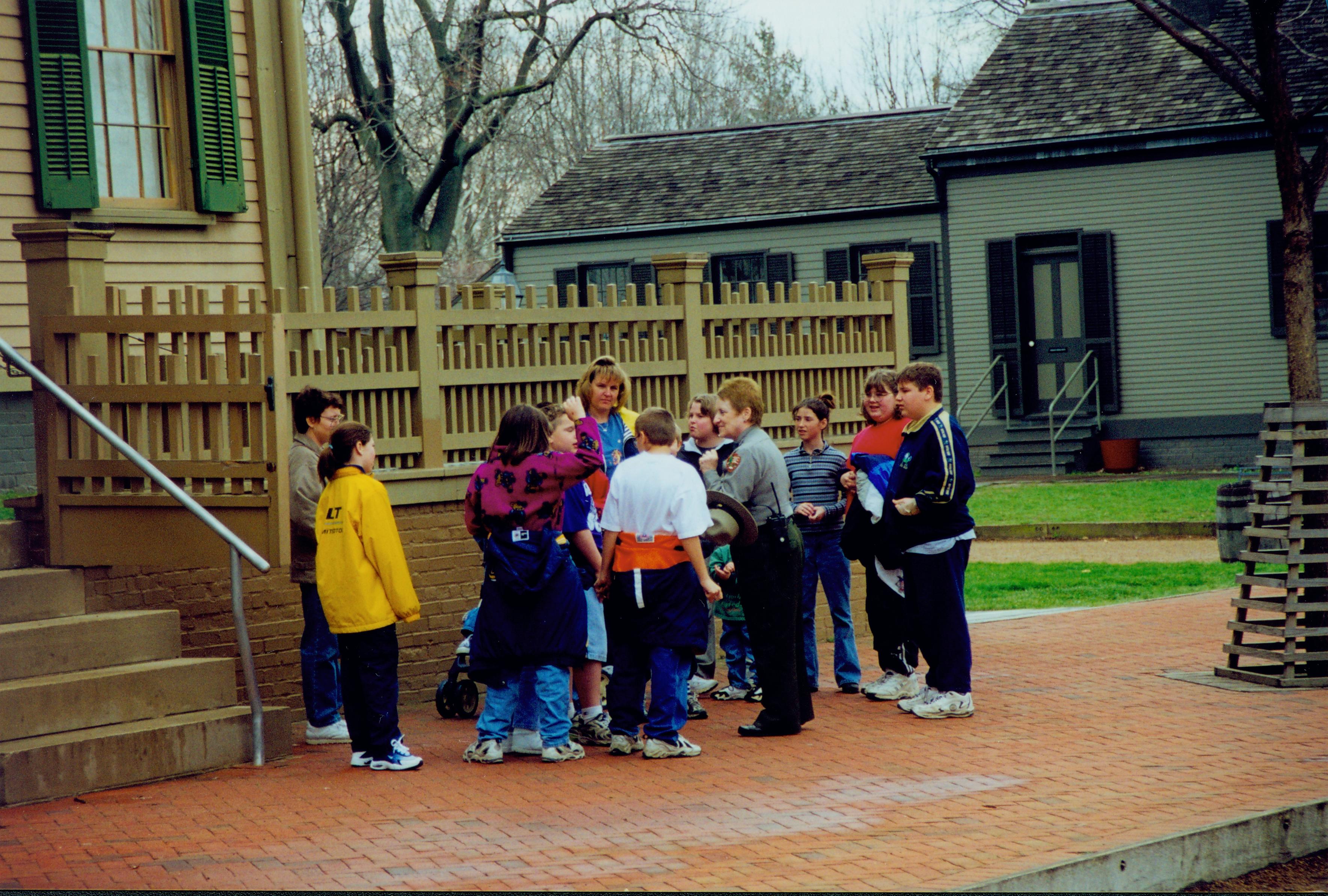 Interpreter Judy Winkelmann speaks with a small group of students in front of the Lincoln Home. The restored Arnold house appears in the background. Photographer facing south east.