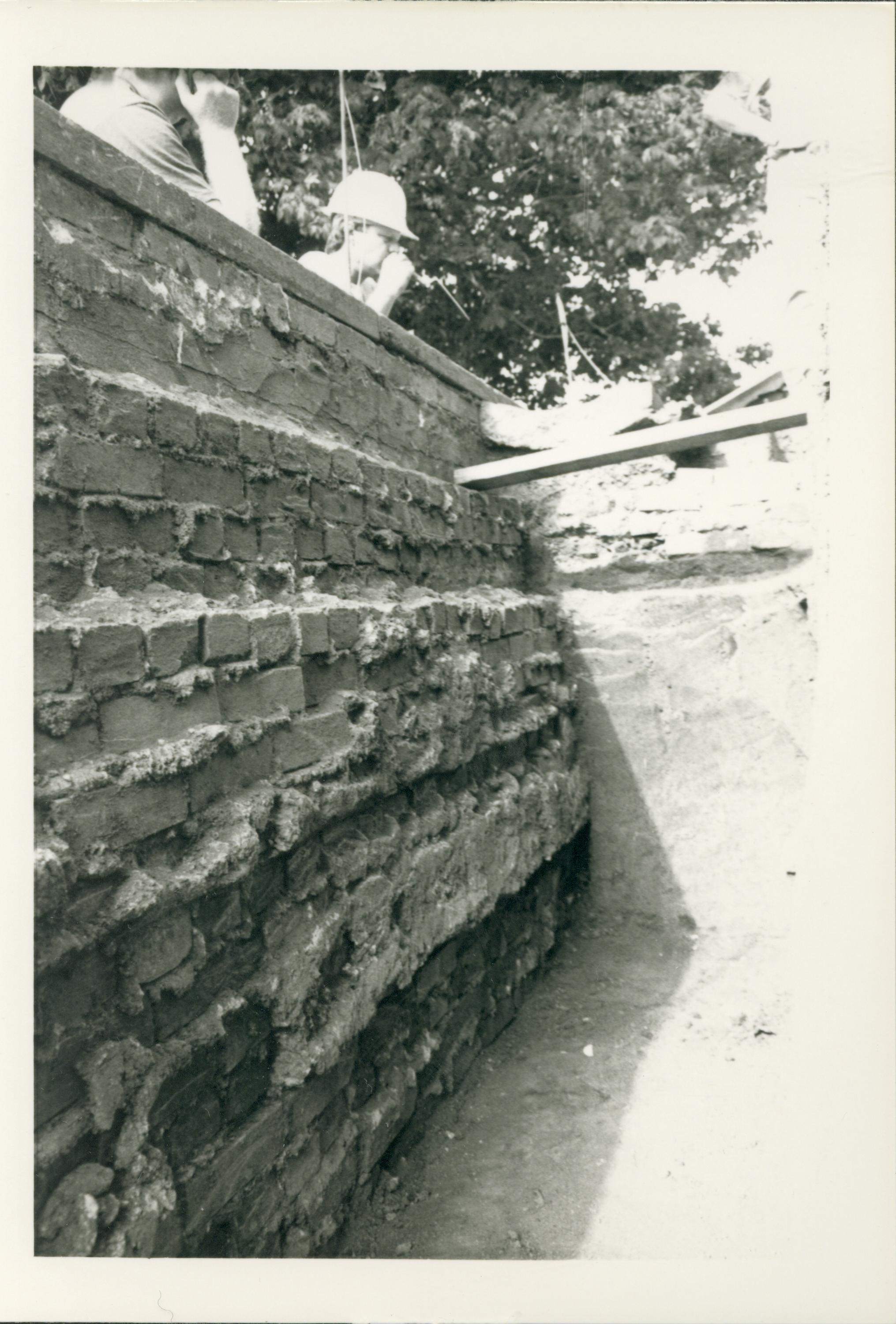 Two workers viewing the trench on the west side of the Lincoln Home during the 1987-1988 restoration. The interior of the west retaining wall can be seen to the left of the picture. The 90 degree turn of the wall to form the north section of the retaining wall can be seen in the middle of the picture. Photographer facing north west.