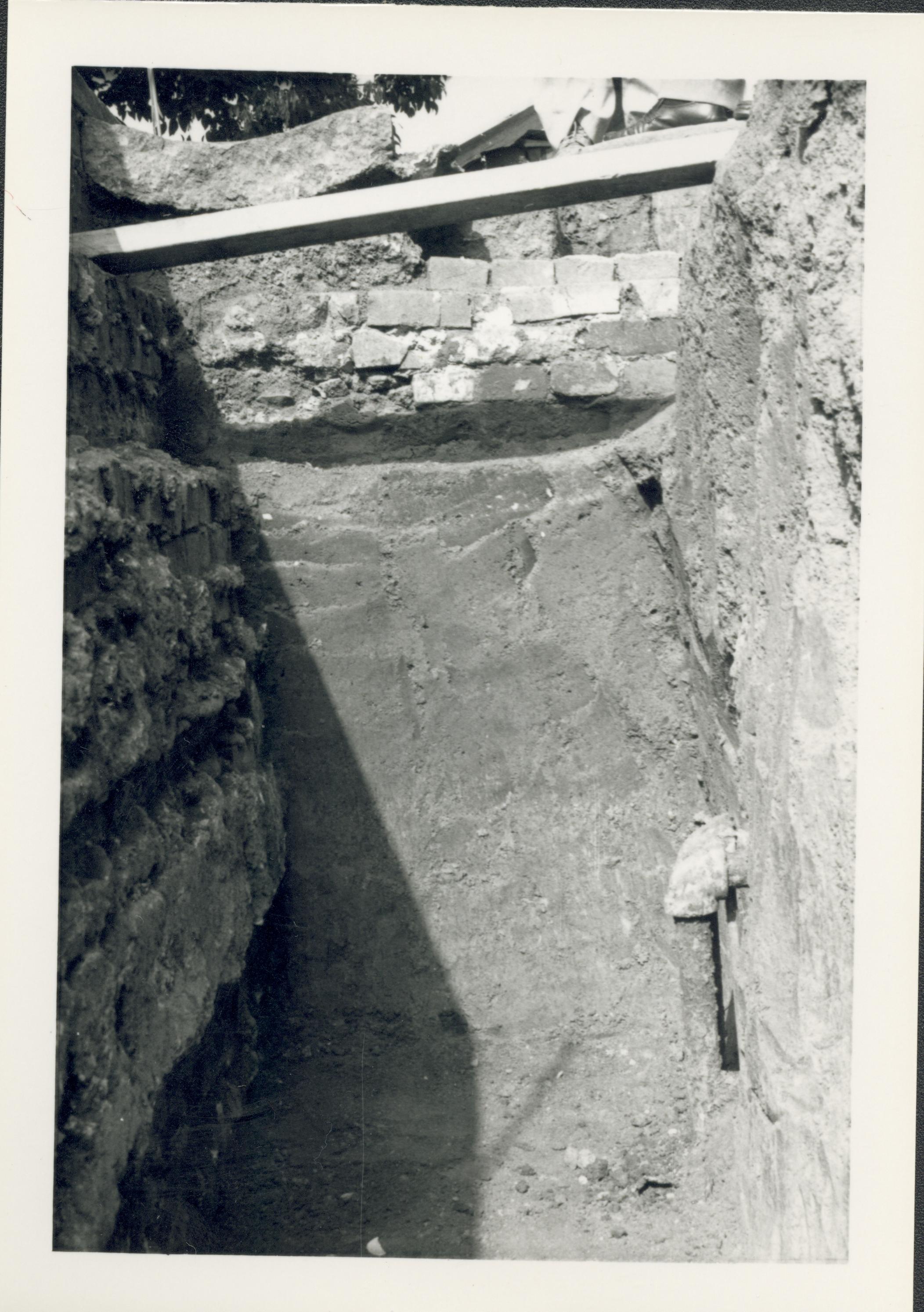 Trench on the west side of the Lincoln Home during the 1987-1988 restoration. The interior of the west retaining wall can be seen to the left of the picture. The 90 degree turn of the wall to form the north section of the retaining wall can be seen in the middle of the picture. There is a pipe exposed on the lower right side of the photo. Photographer facing north.