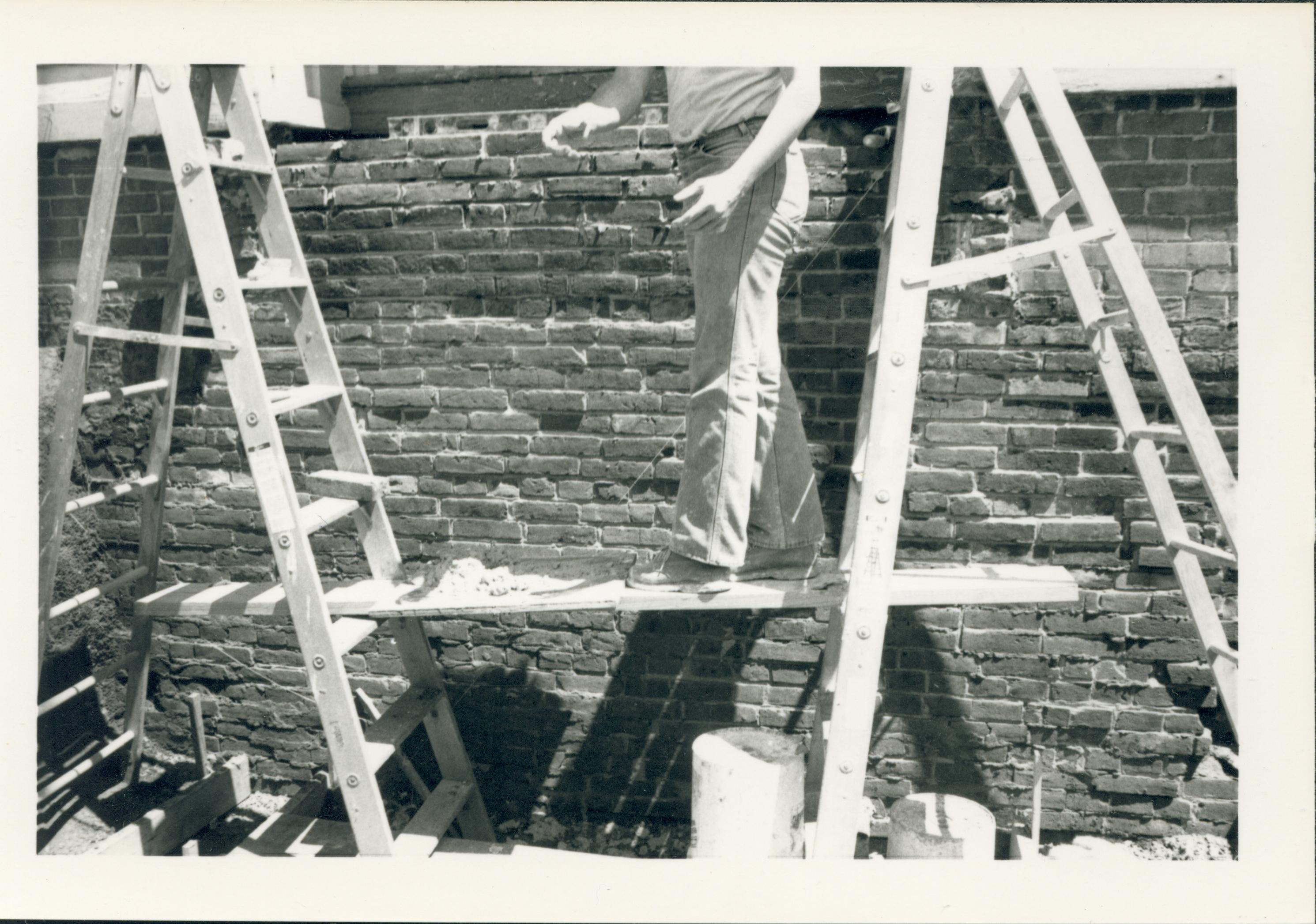 Two ladders in a trench alongside an exposed section of the Lincoln Home foundation during the 1987-1988 restoration. A single worker is standing on a platform supported by the two ladders. The west entrance to the Lincoln Home can be seen at top.