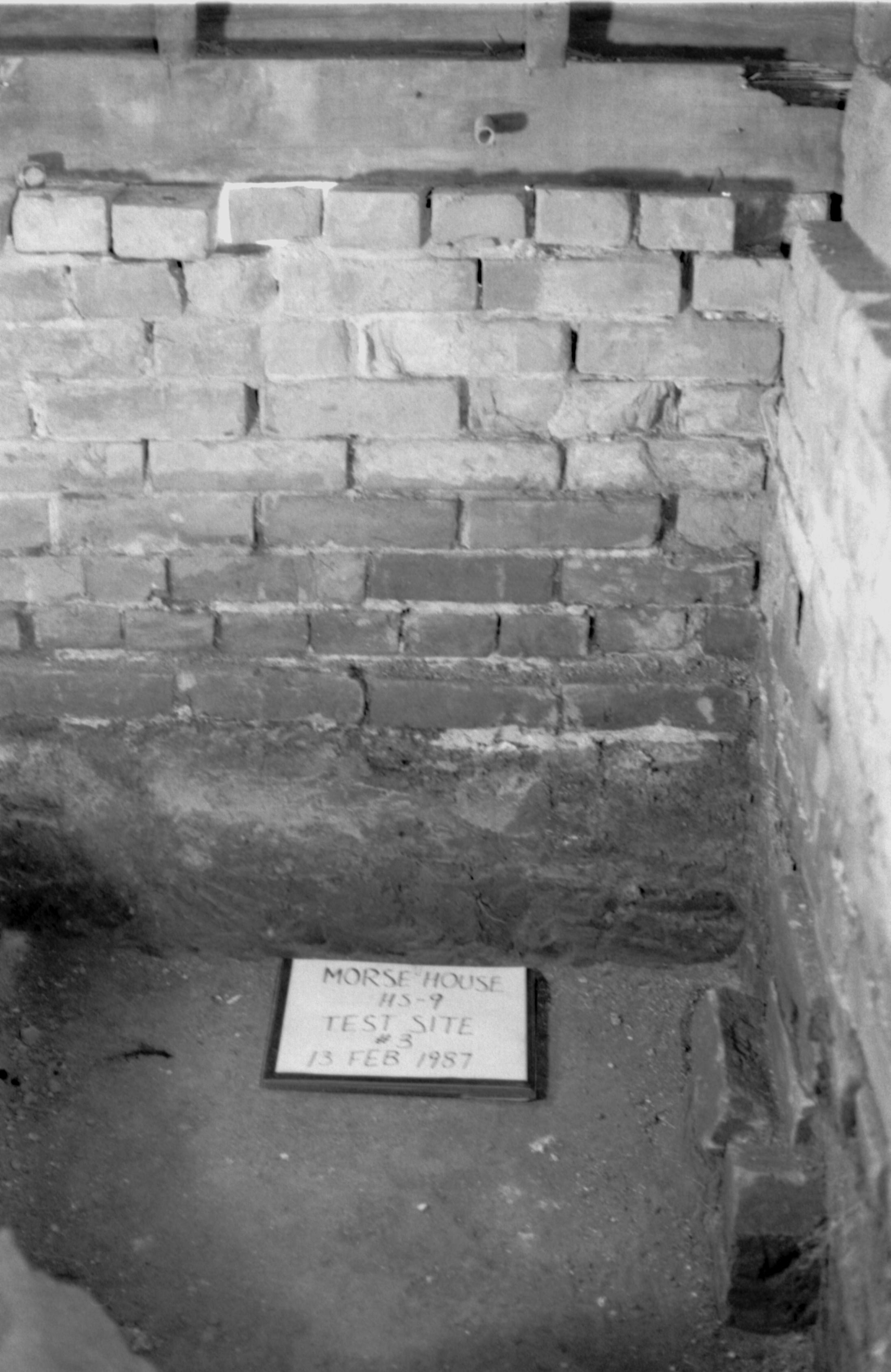 Morse House HS-9 Test Site #3. Corner of basement, showing intersection of brick foundation, sill, studs, and sheathing. Test pit is in foreground, with id plaque.
