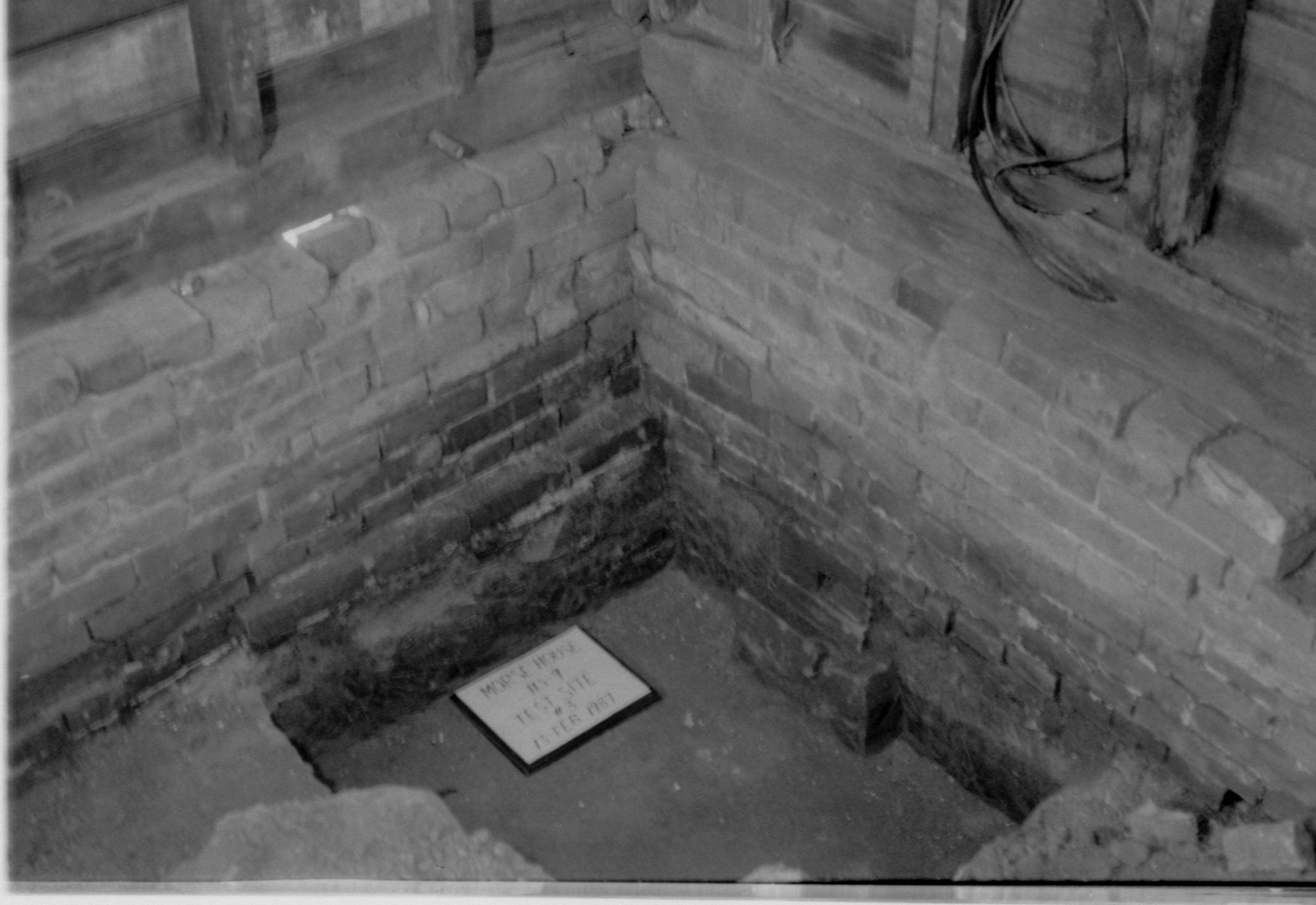 Morse House HS-9 Test Site #3. Corner of basement, showing intersection of brick foundation, sill, studs, and sheathing. Test pit is in foreground, with id plaque.